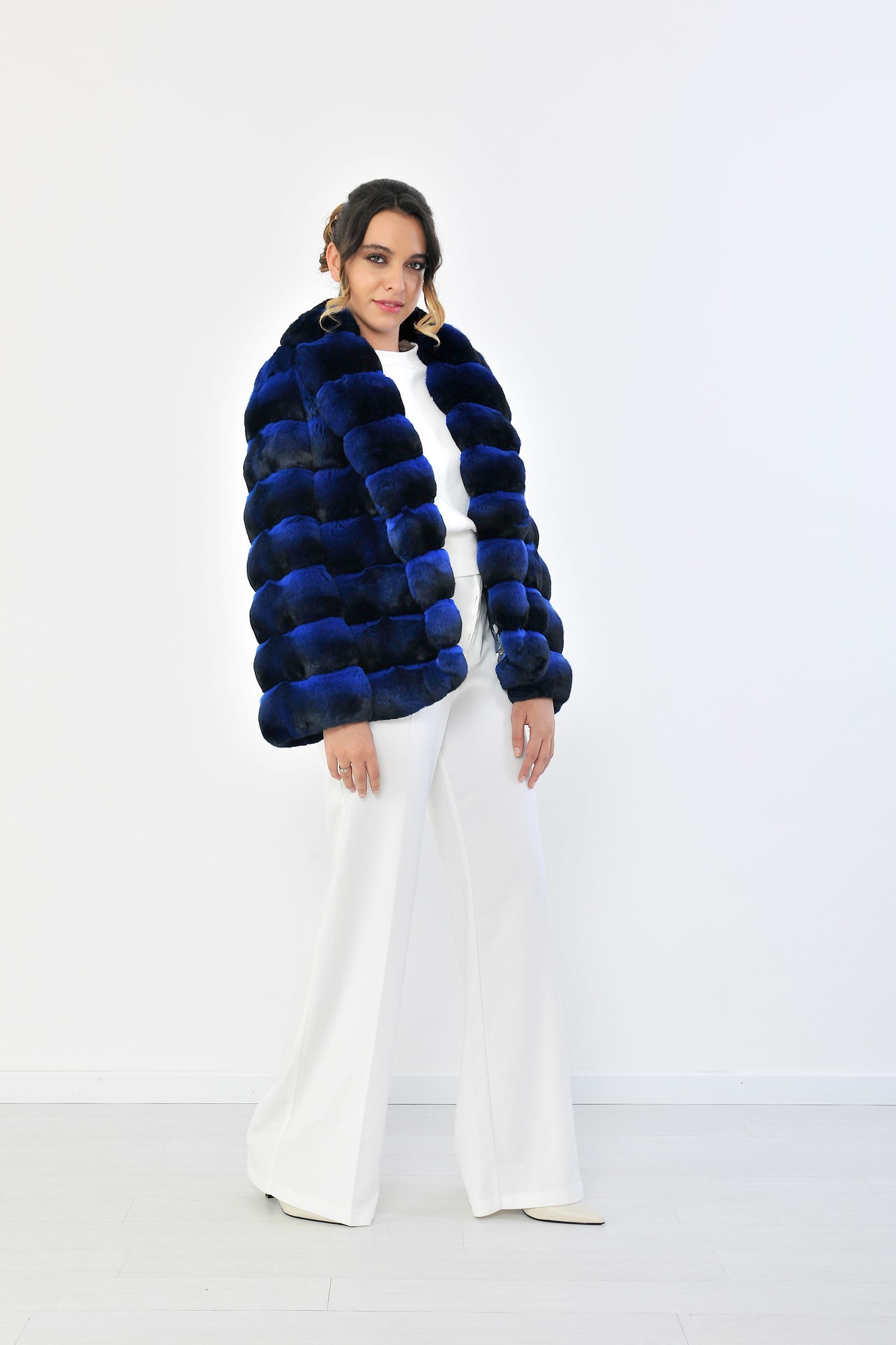 Una donna con una giacca di pelliccia blu e pantaloni bianchi è in piedi su uno sfondo bianco.