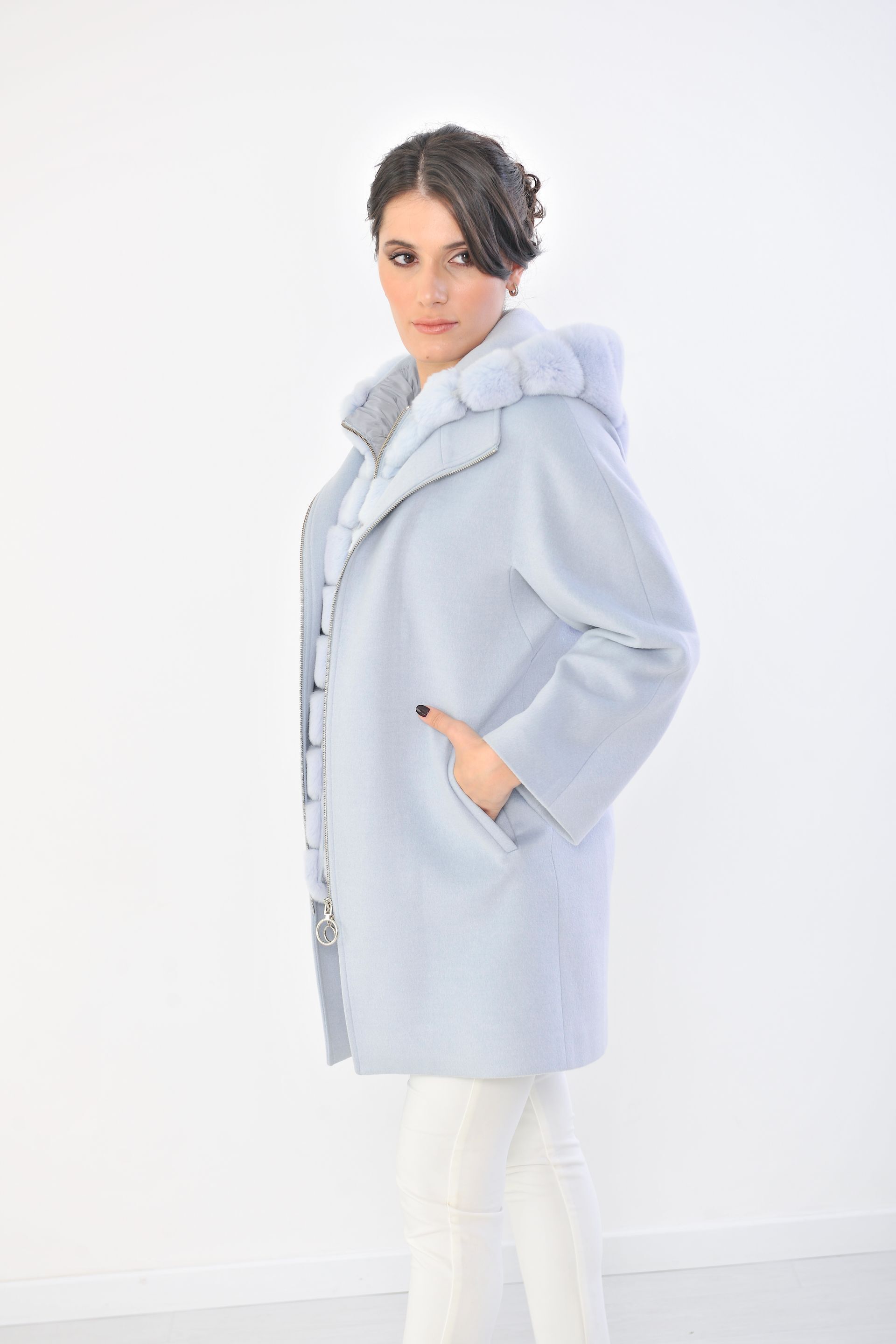 Donna che indossa un cappotto azzurro con rifiniture in pelliccia, pantaloni bianchi, mano in tasca, su sfondo bianco.