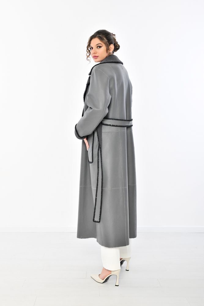 Donna con lungo cappotto grigio con rifiniture nere, in piedi di lato, pantaloni bianchi, tacchi.
