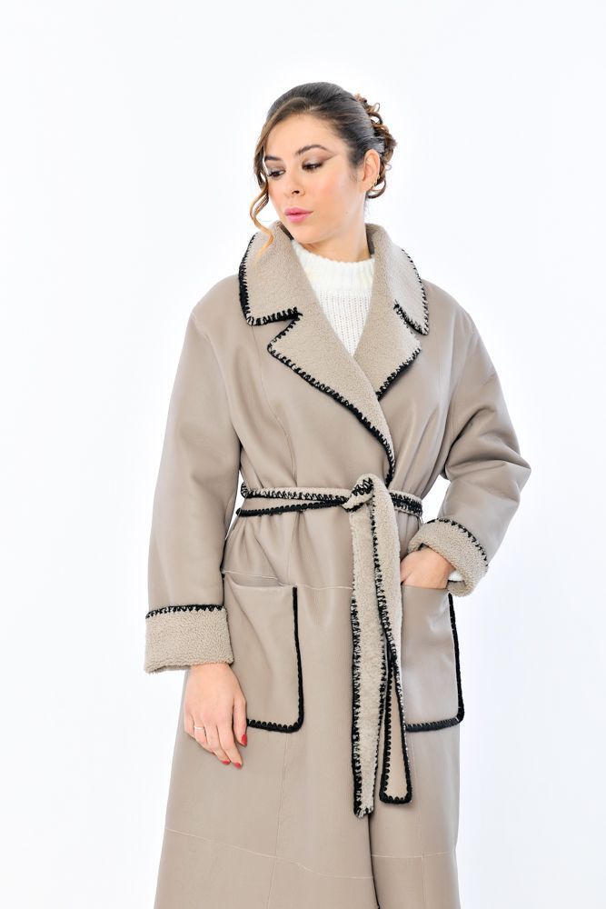 Donna in piedi con cappotto beige con rifiniture e cintura nere e maglione bianco.