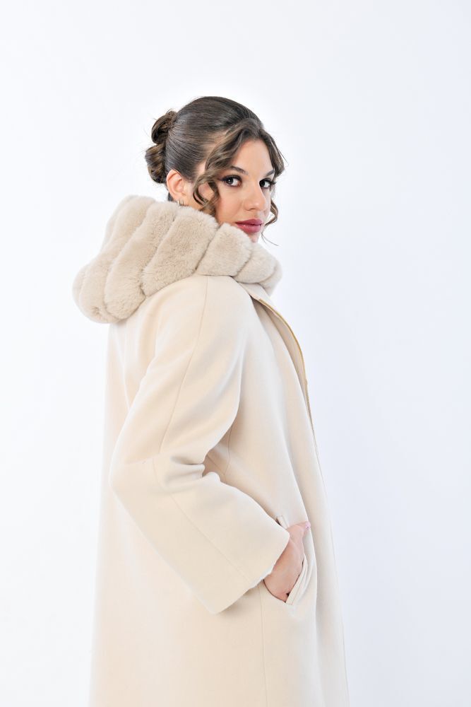 Donna con cappotto color crema e cappuccio bordato di pelliccia, che guarda oltre le spalle, su sfondo bianco.