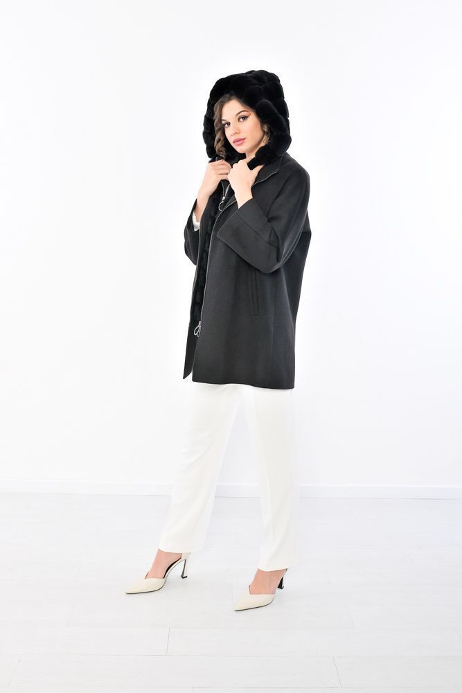 Donna con cappotto nero, cappuccio di pelliccia e pantaloni bianchi, in piedi su uno sfondo bianco.