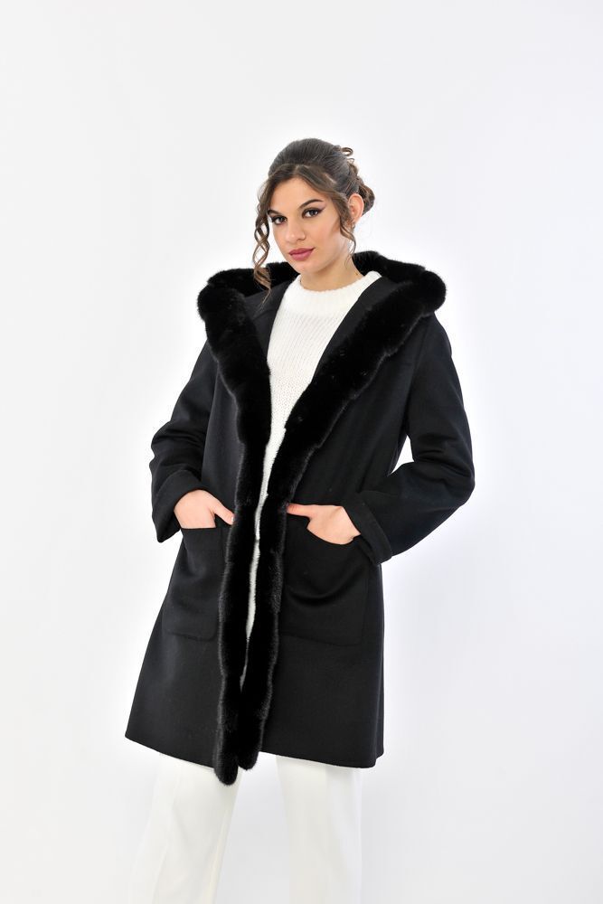 Donna con cappotto nero e cappuccio foderato di pelliccia, mani in tasca, su sfondo bianco.