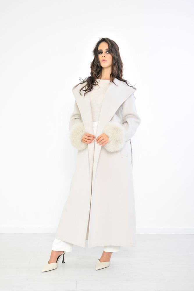Donna con un lungo cappotto chiaro con polsini di pelliccia, in piedi in un interno su uno sfondo bianco.