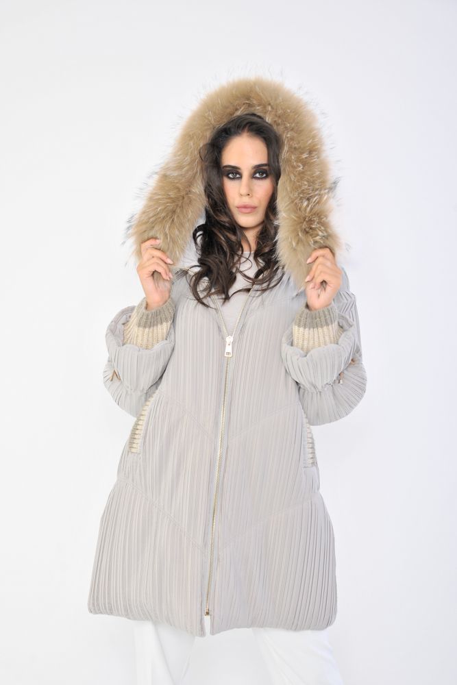 Donna con cappotto grigio chiaro e cappuccio foderato di pelliccia, mani alzate, in piedi davanti a uno sfondo bianco.