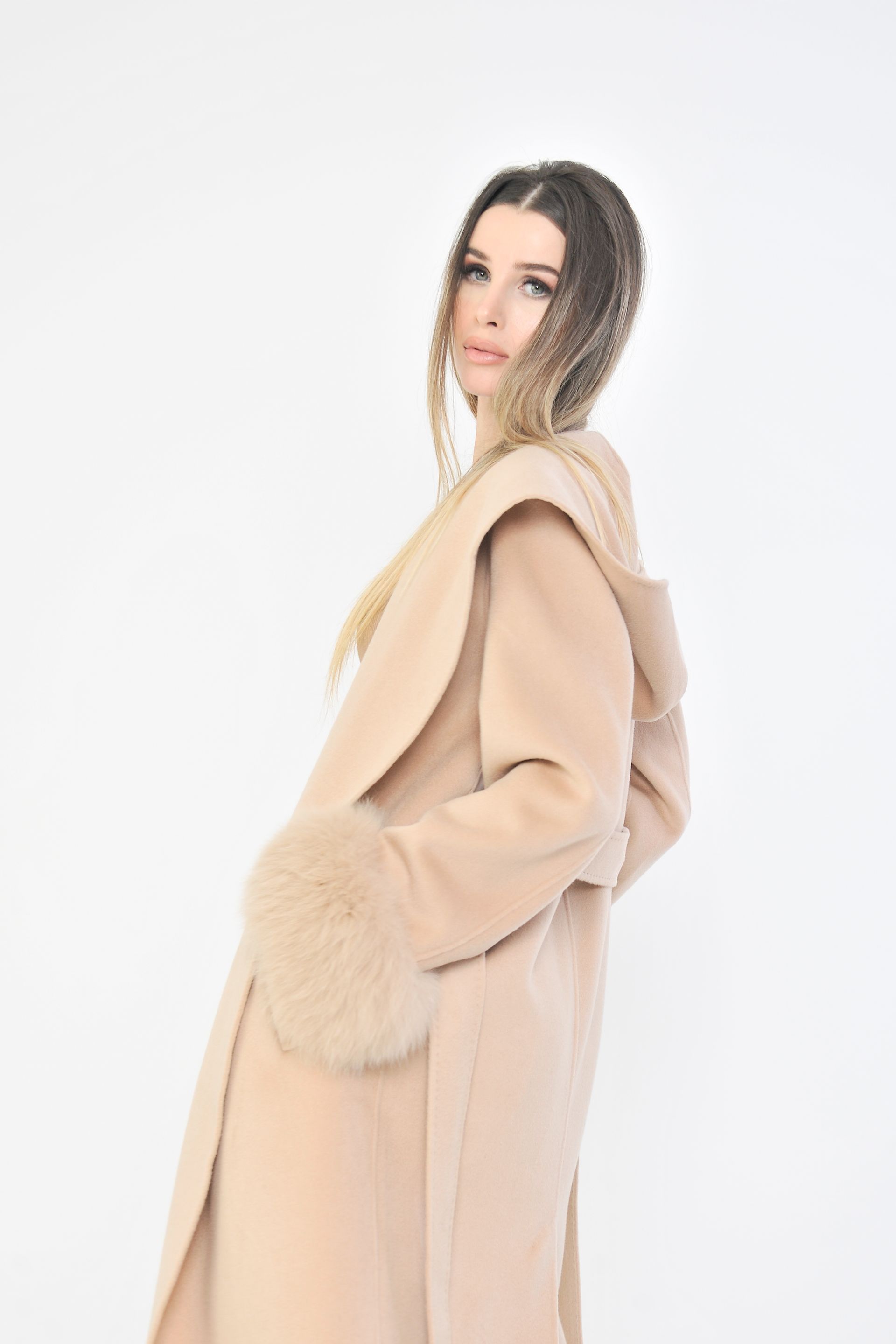 Donna con cappotto marrone chiaro con polsini di pelliccia, cappuccio alzato, in posa davanti a uno sfondo bianco.