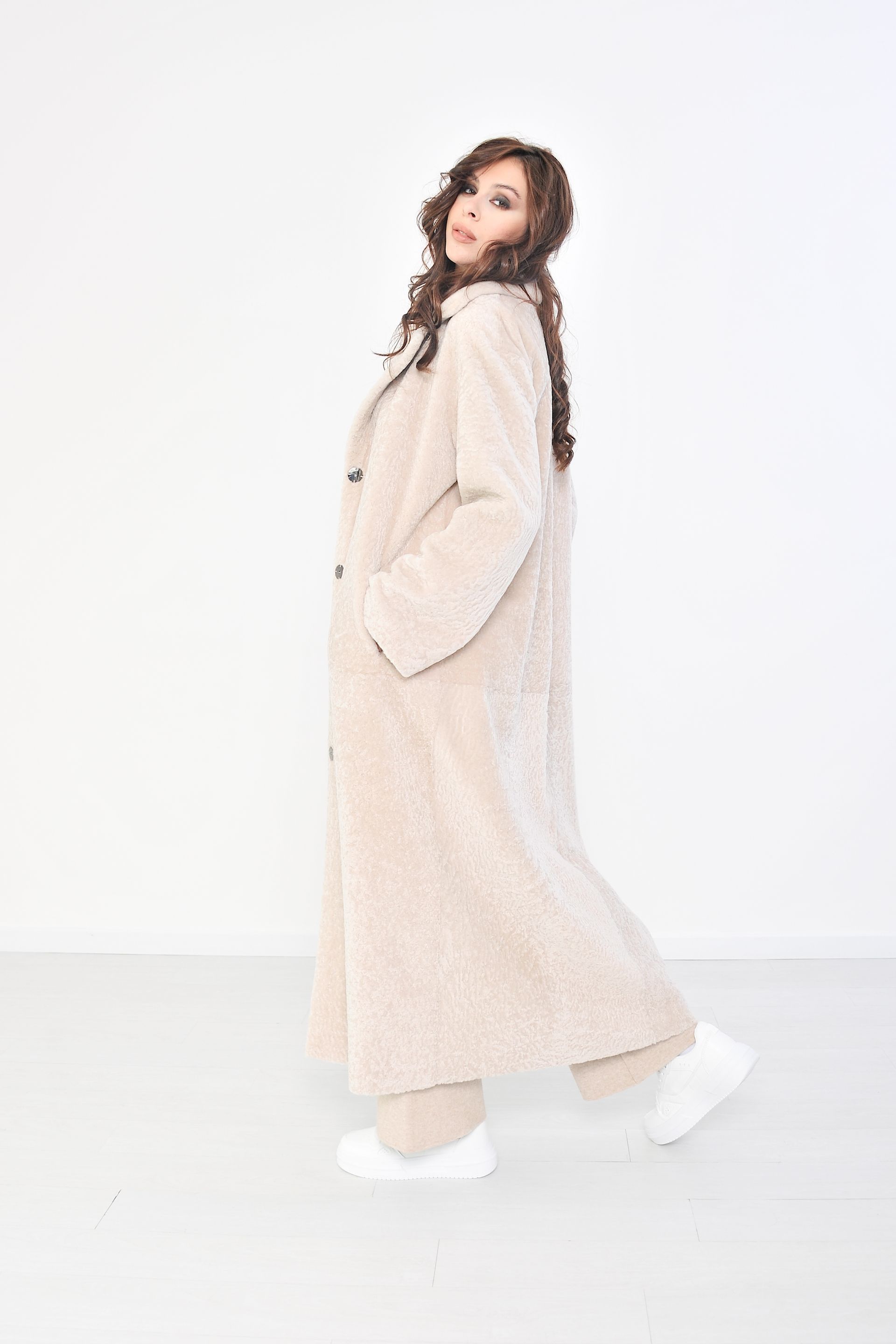 Donna che indossa un lungo cappotto beige e pantaloni, in piedi in una stanza bianca.