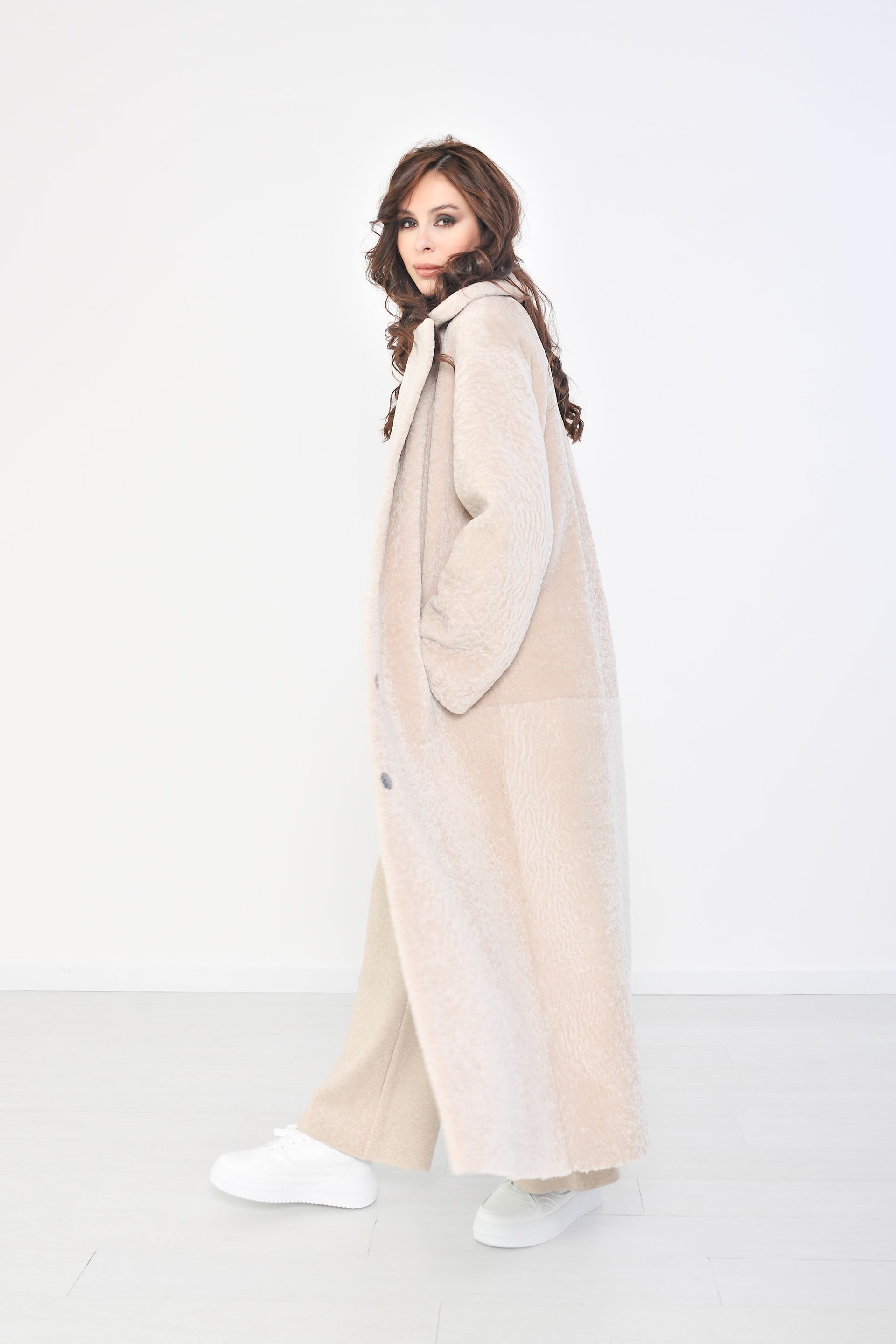 Donna che indossa un lungo cappotto beige e pantaloni, in piedi in una stanza bianca.