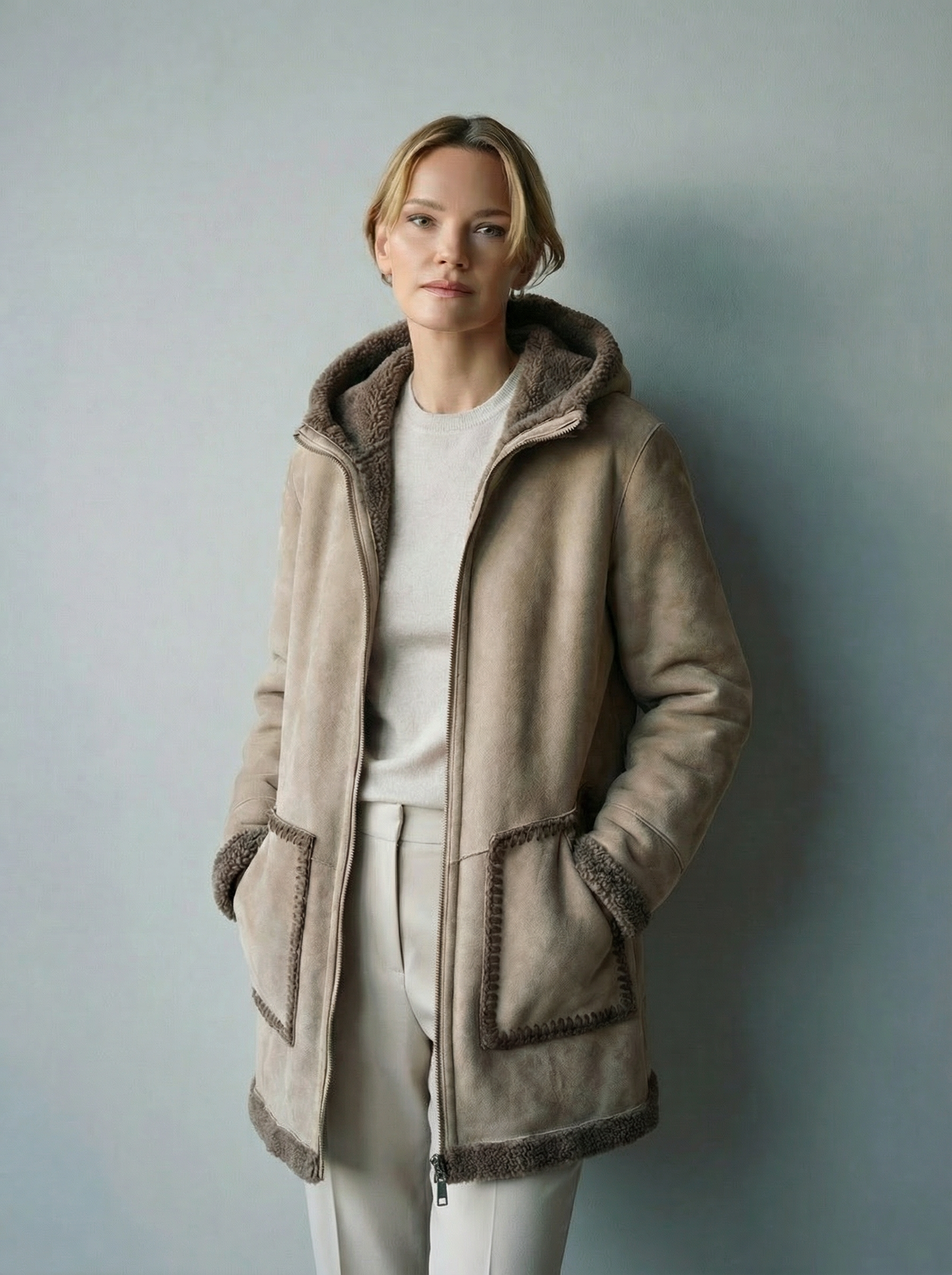 Donna in cappotto beige con cappuccio e tasche, in piedi in casa.
