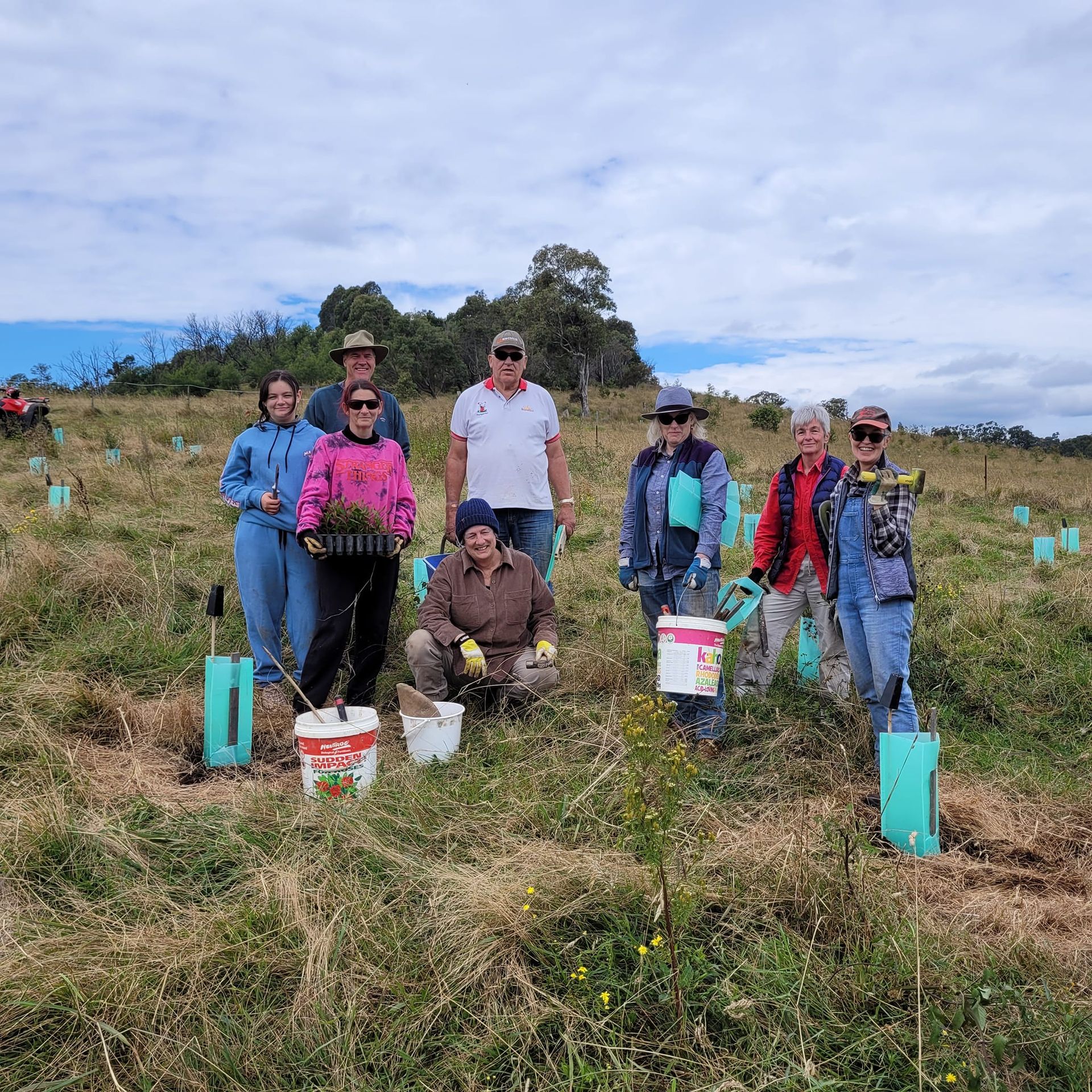 Group of people standing in a grassy field with tree seedlings for planting.