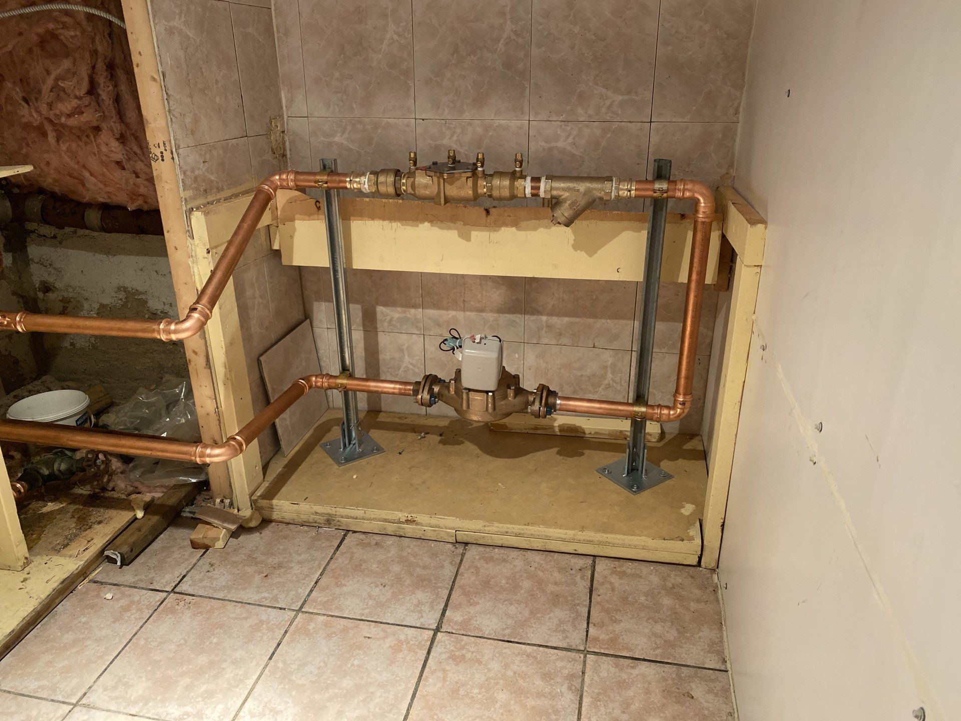 Copper pipes and valves installed in a tiled alcove, resting on wooden framing and supported by metal legs.