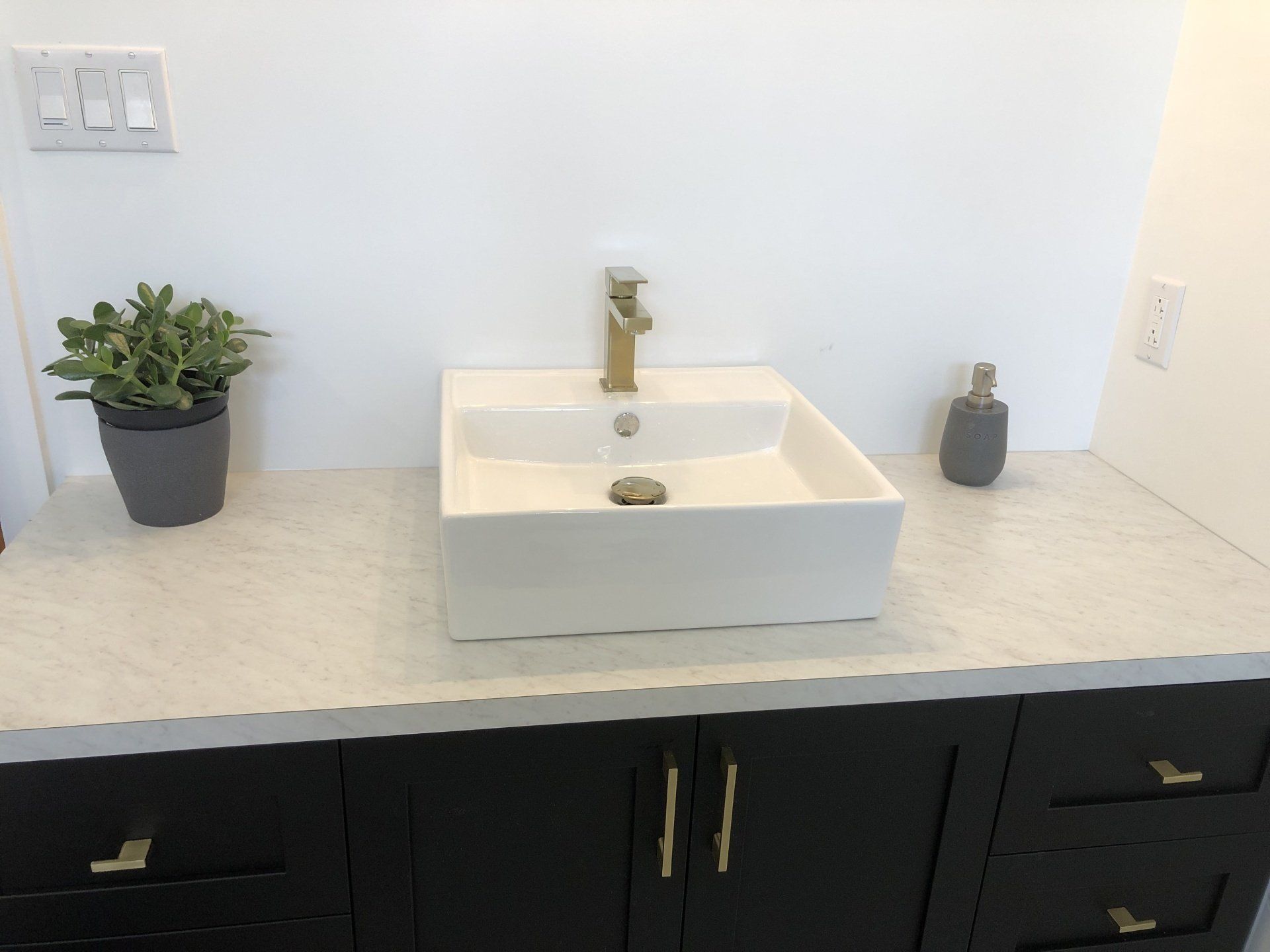 White countertop with black cabinets, a square white sink, and gold faucet.