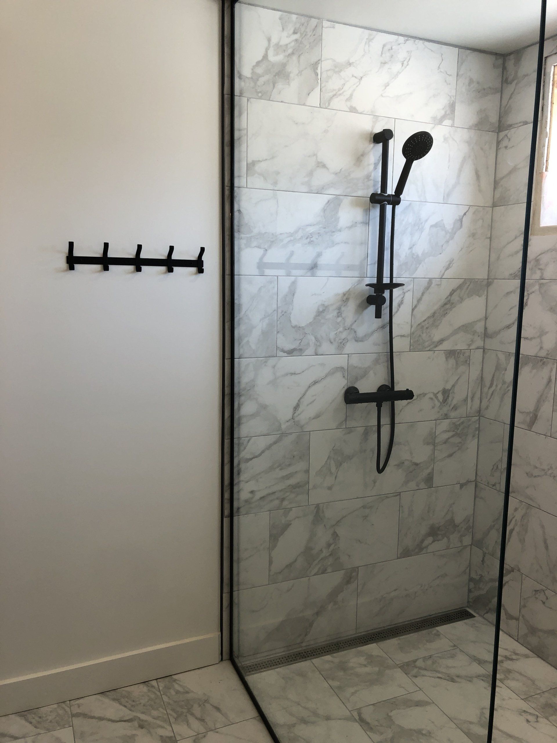 Shower with marble-look tile, black fixtures, and a glass door. A wall hook is to the left.