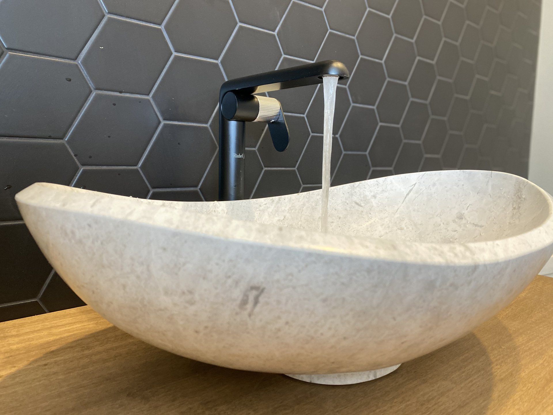 Water flows from a black faucet into a white, stone vessel sink. Hexagonal dark gray tiles form the backdrop.