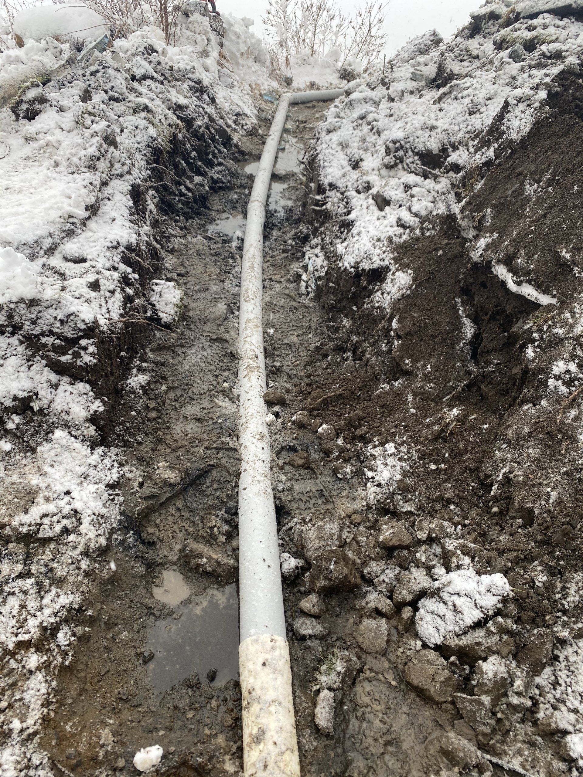 Un tuyau blanc posé dans une tranchée étroite et boueuse recouverte de neige.