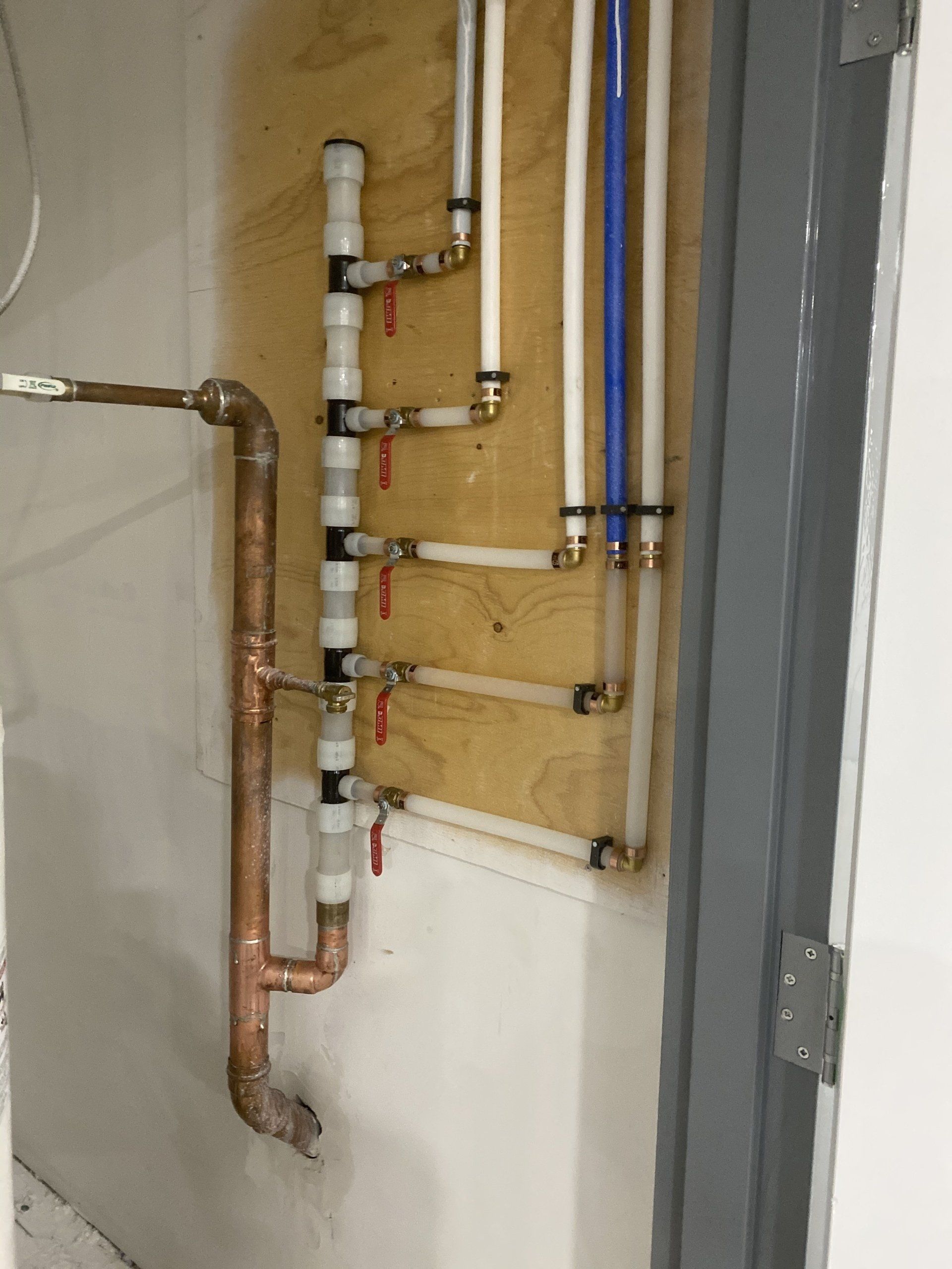 Plumbing pipes of various types installed against a wooden wall, including copper and white/blue plastic.