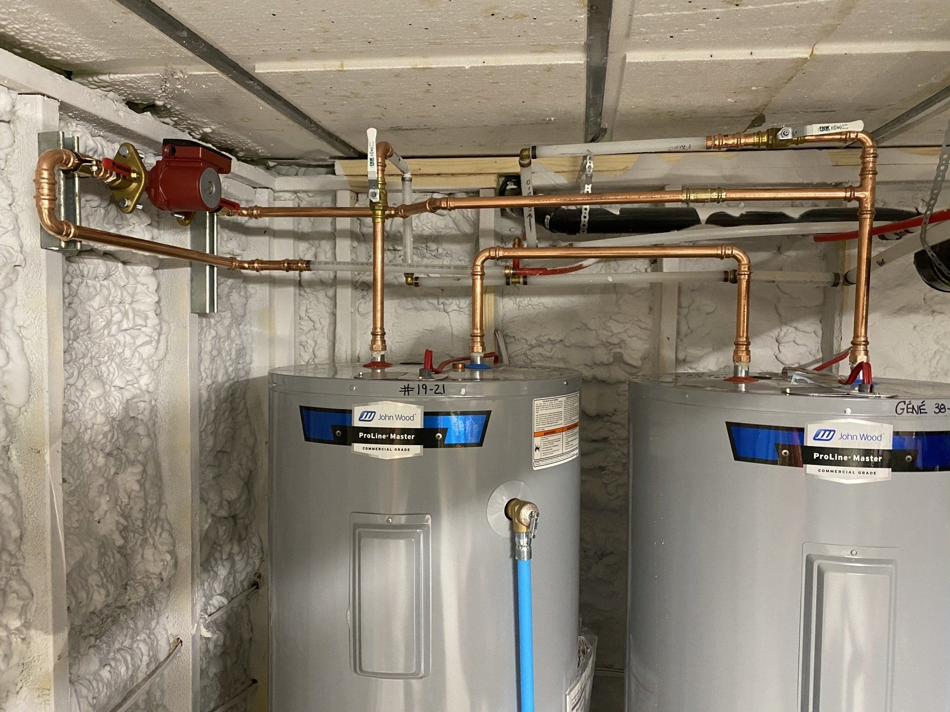 Two water heaters with copper pipes and a red pump in a concrete-walled room.