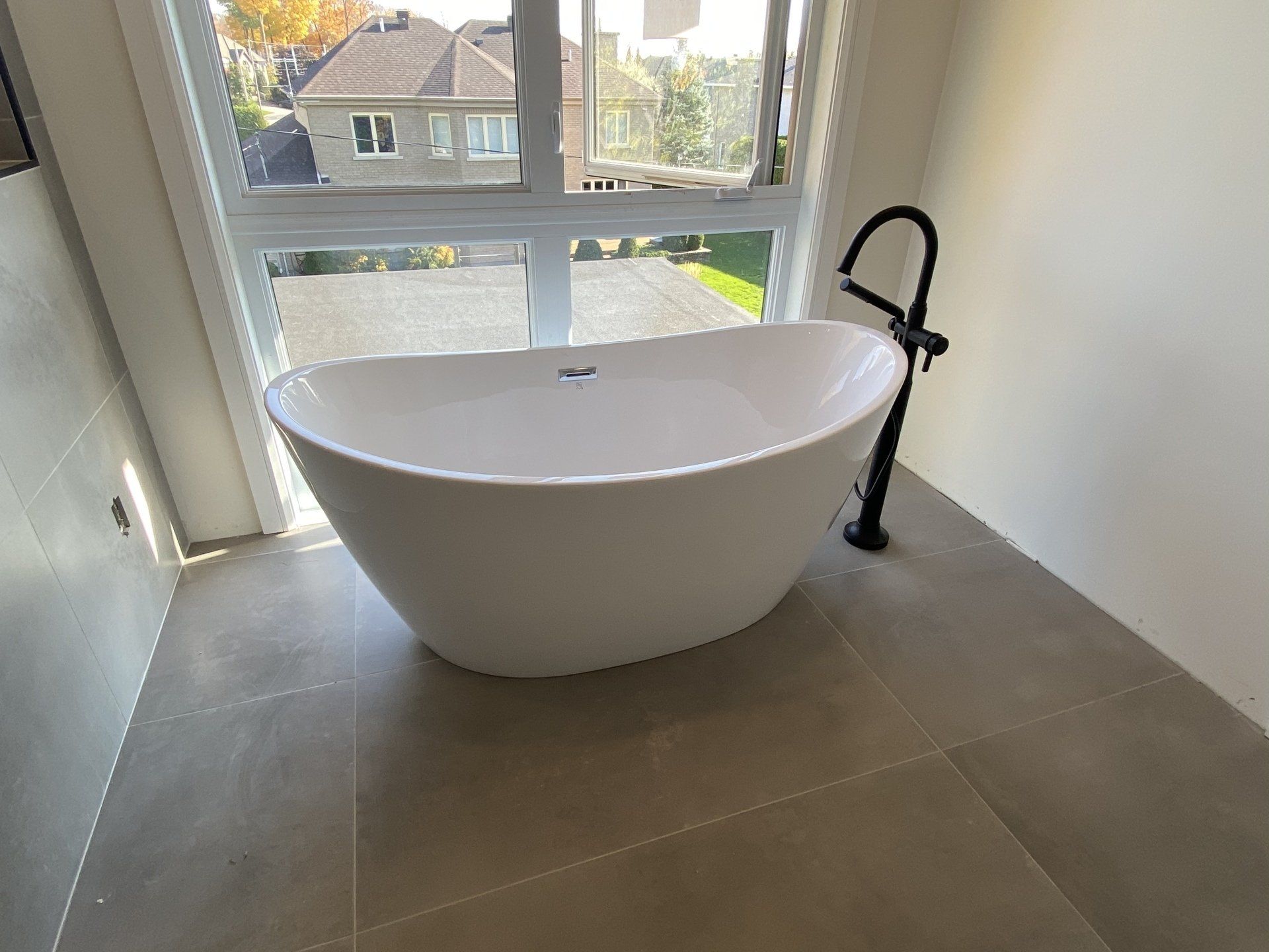 White freestanding bathtub near window with black faucet; gray tiled floor.