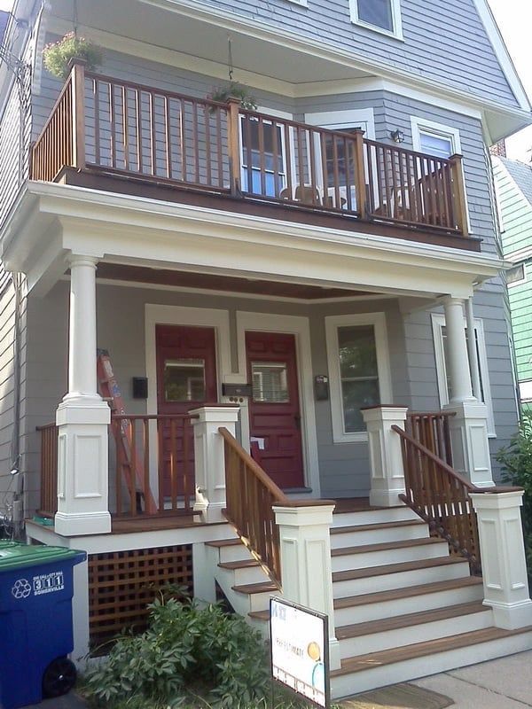 Two-story house with front porch and balcony. Gray siding, brown railing, white columns. Red doors. Blue trash bin on left.