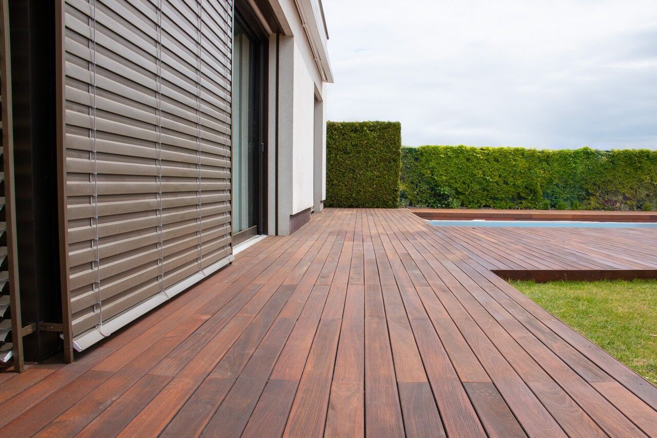 Wooden deck extending from a house with closed brown shutters, leading to a pool and lawn.