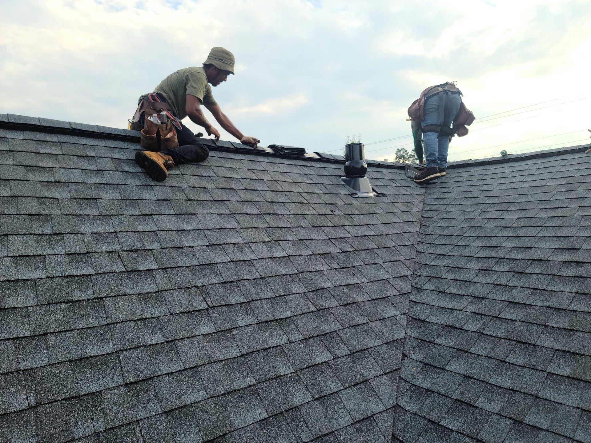 Roofer installing dark gray tiles on a partially completed roof with a red drill; sunny daylight.