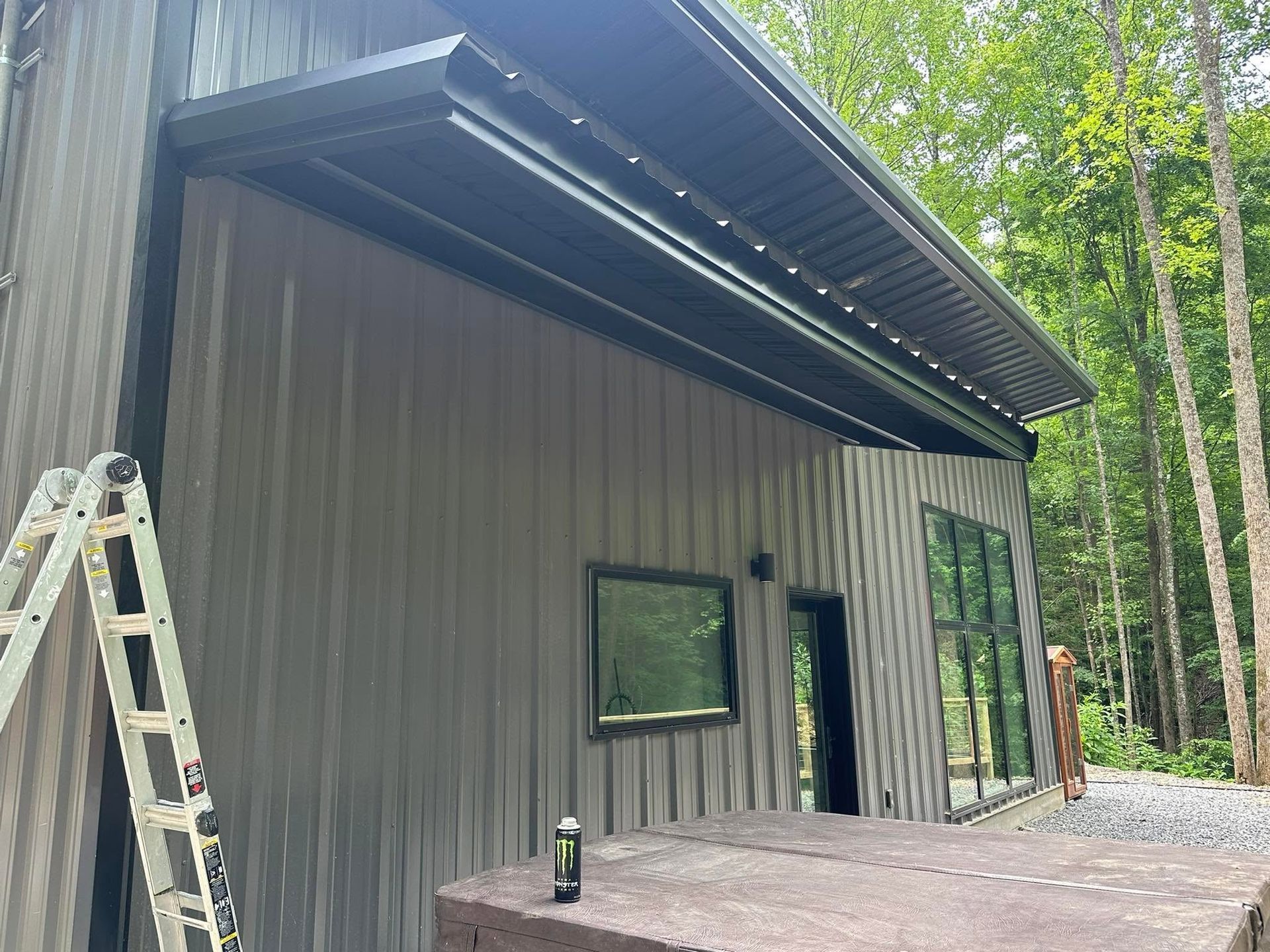 Gray metal building with a covered overhang, windows, and a ladder beside it in a wooded setting
