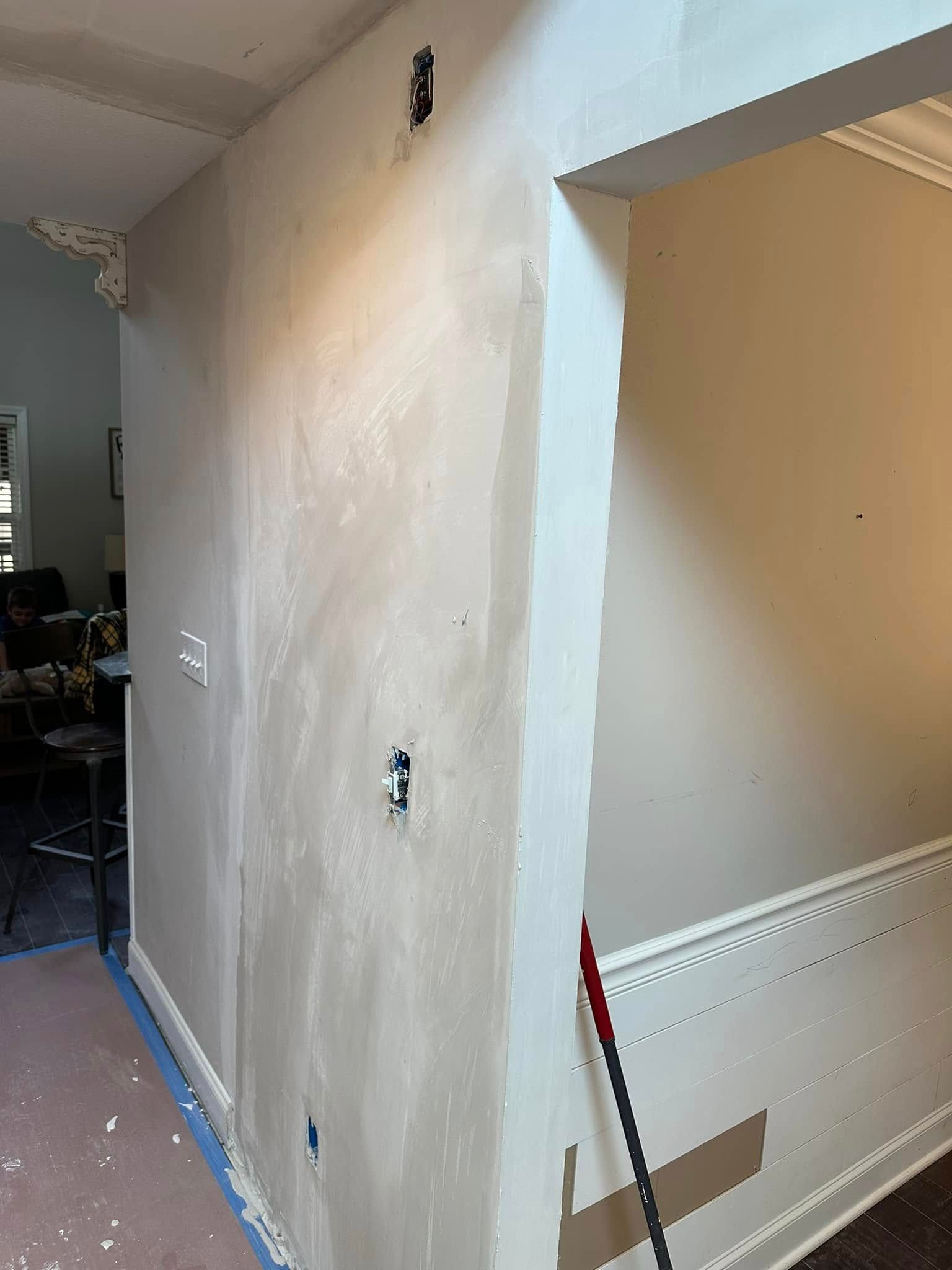 Partially painted hallway wall with patchy white primer, blue tape, and a broom leaning against it
