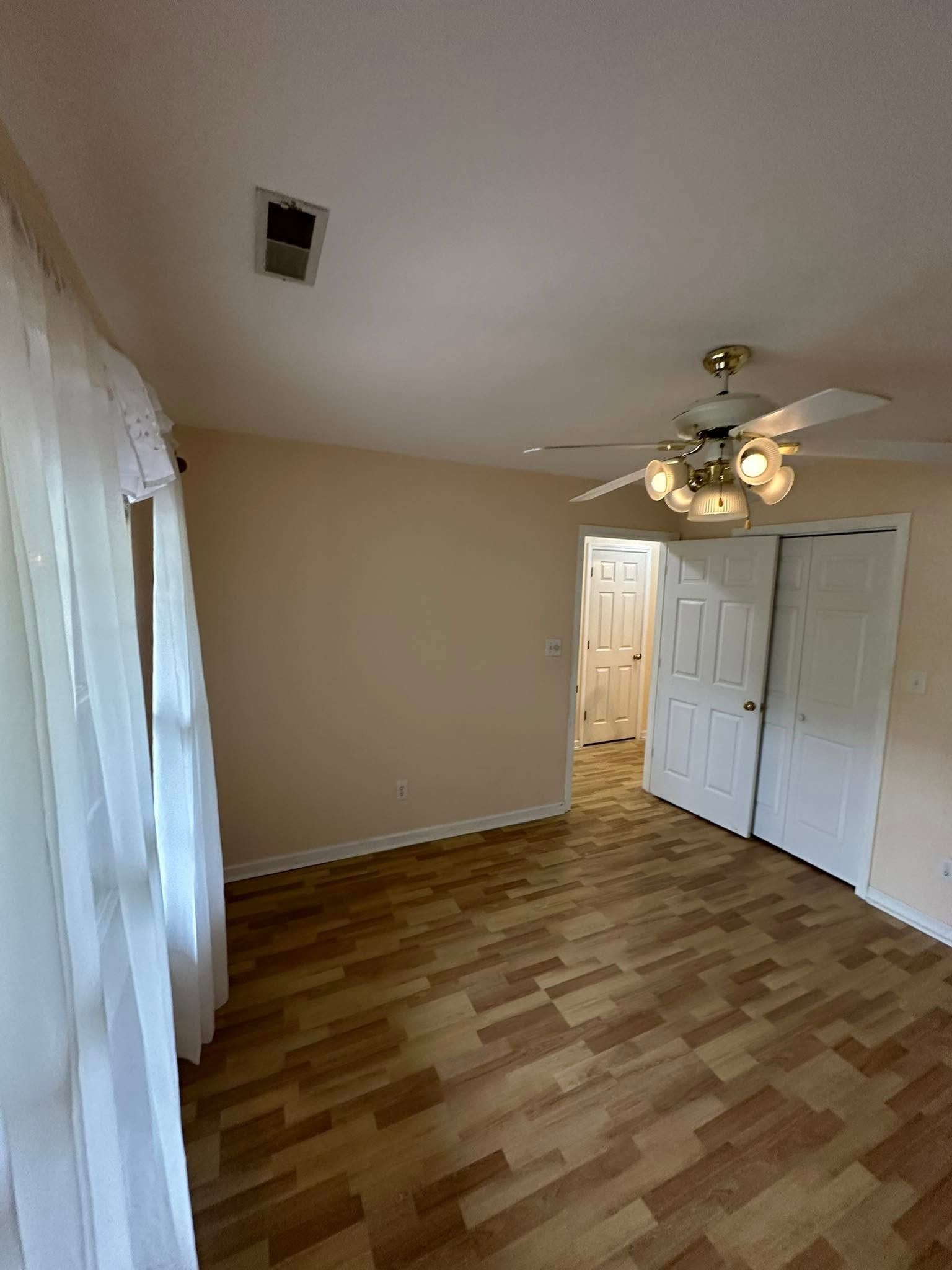 Empty beige room with wood-pattern floor, ceiling fan, and white closet doors near a doorway.