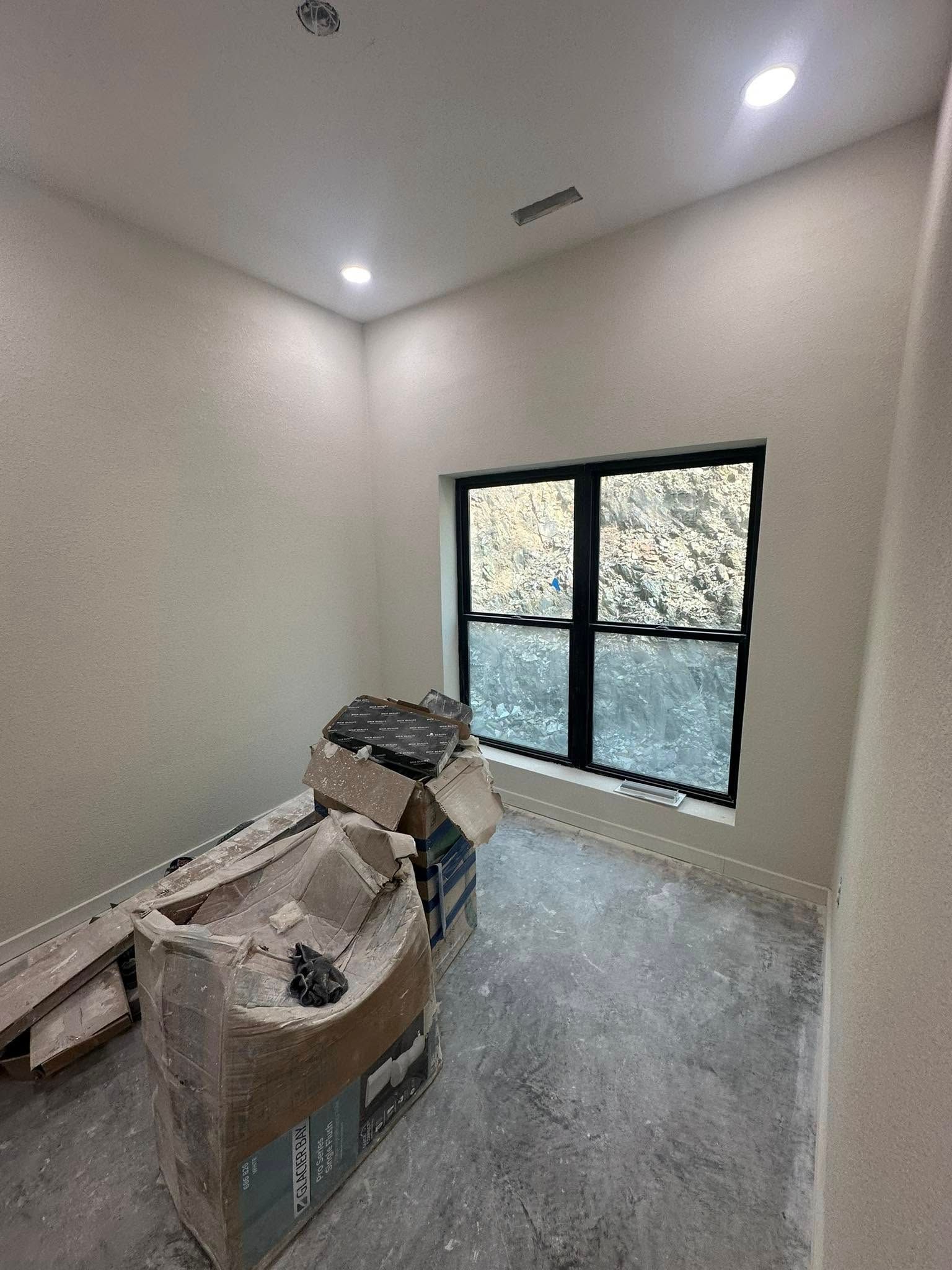 Small empty room with beige walls, recessed lights, a window, and stacked construction materials on the floor.