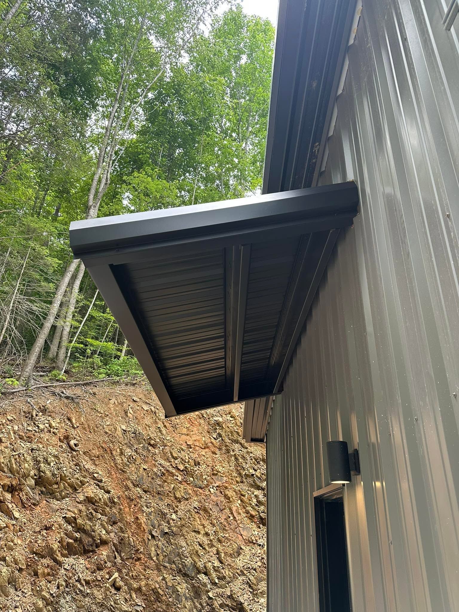 Black metal awning attached to a gray building beside a wooded hillside