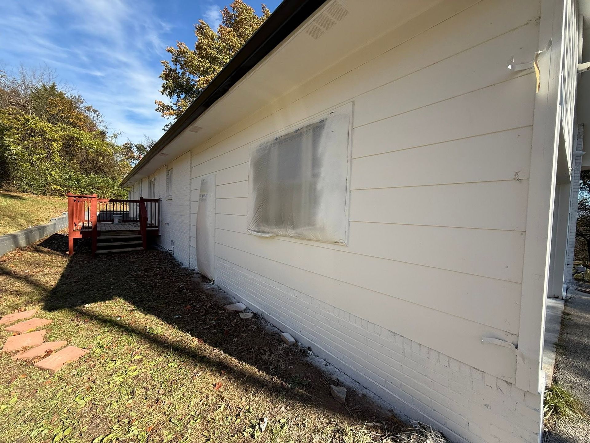 White house exterior with a small window, red deck, and sloping yard in sunlight
