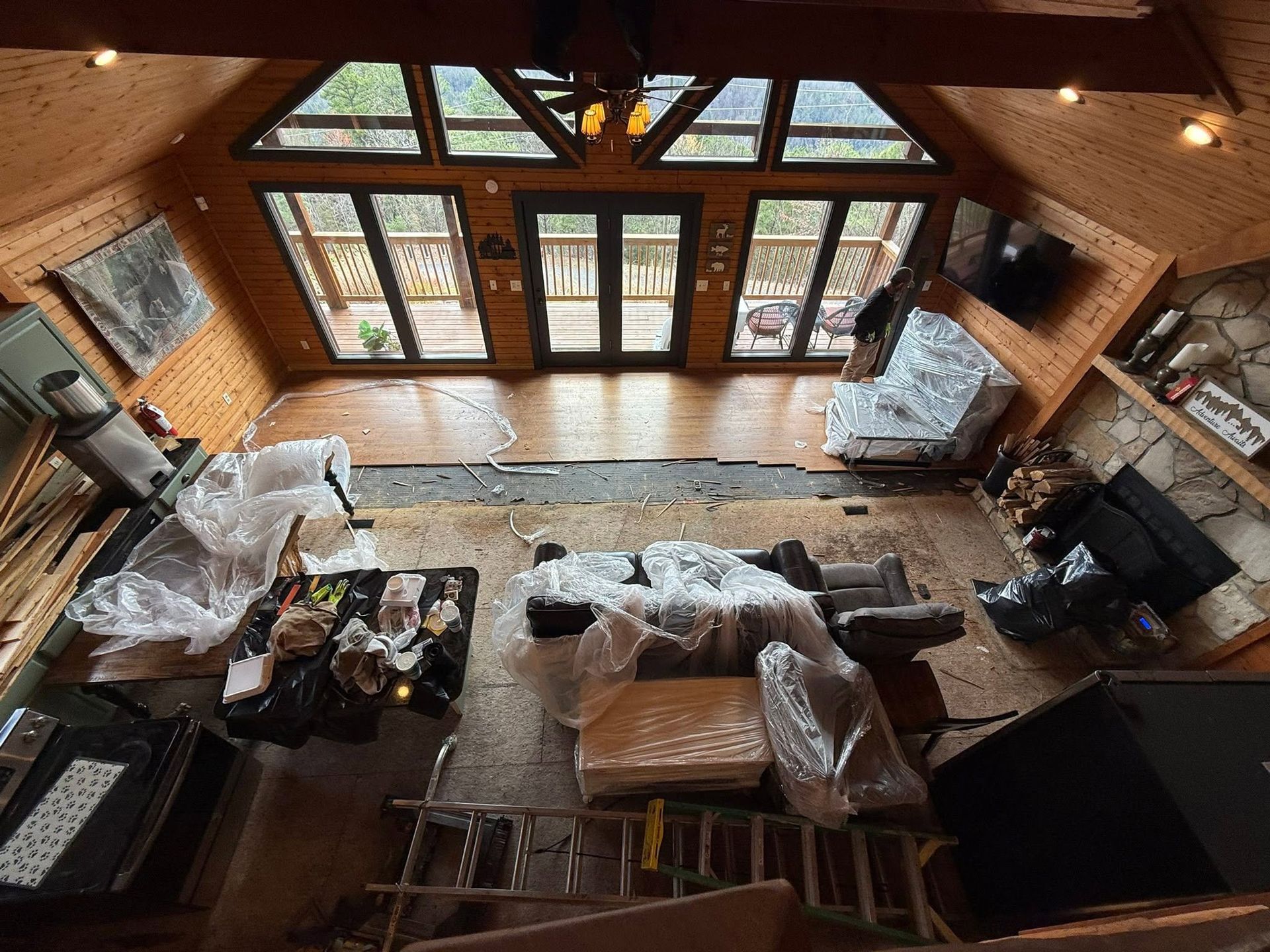Wood-paneled cabin living room with large windows, furniture covered in plastic, and a stone fireplace.