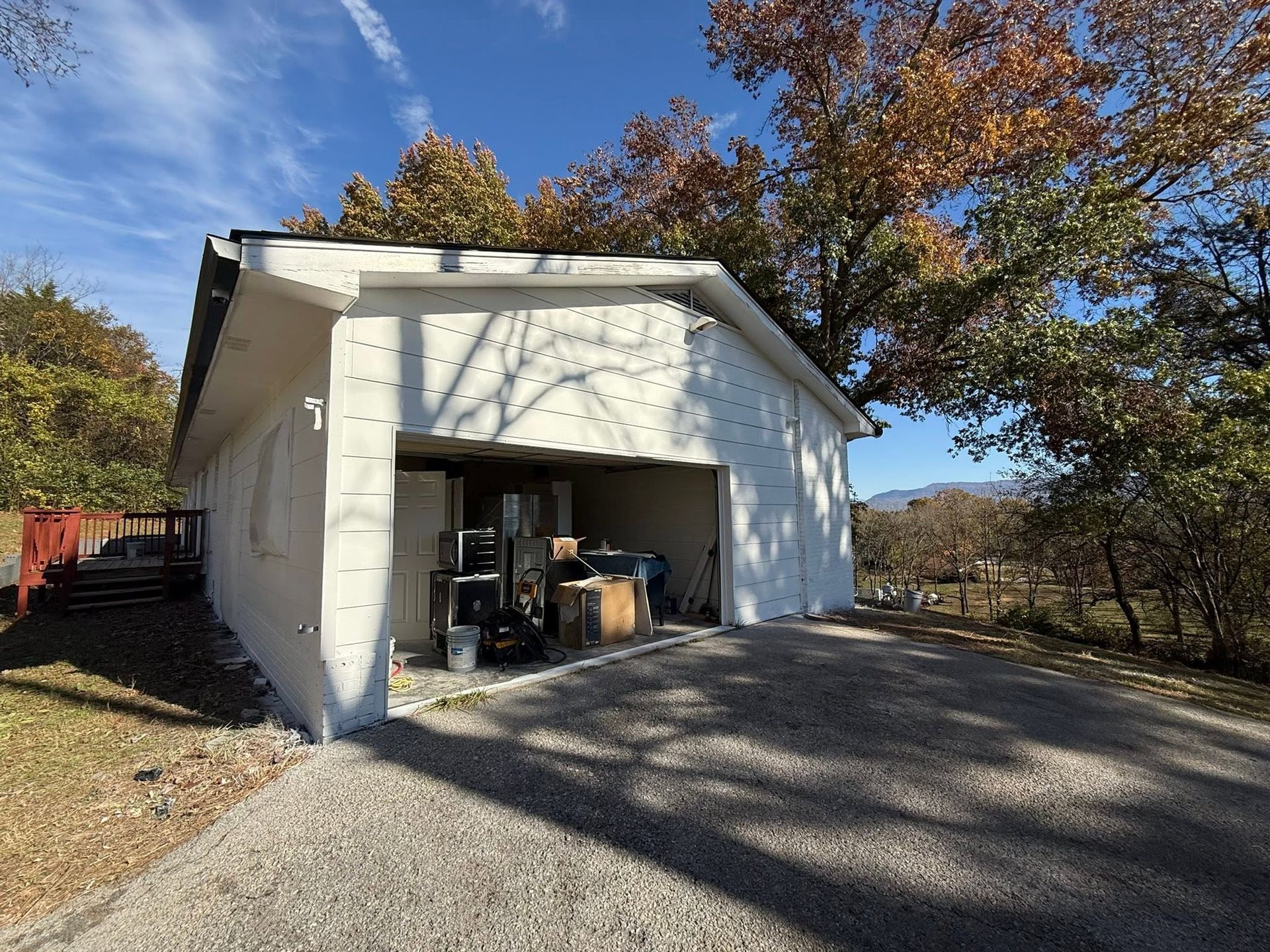 White garage with open bay on a gravel driveway, surrounded by autumn trees under a blue sky.