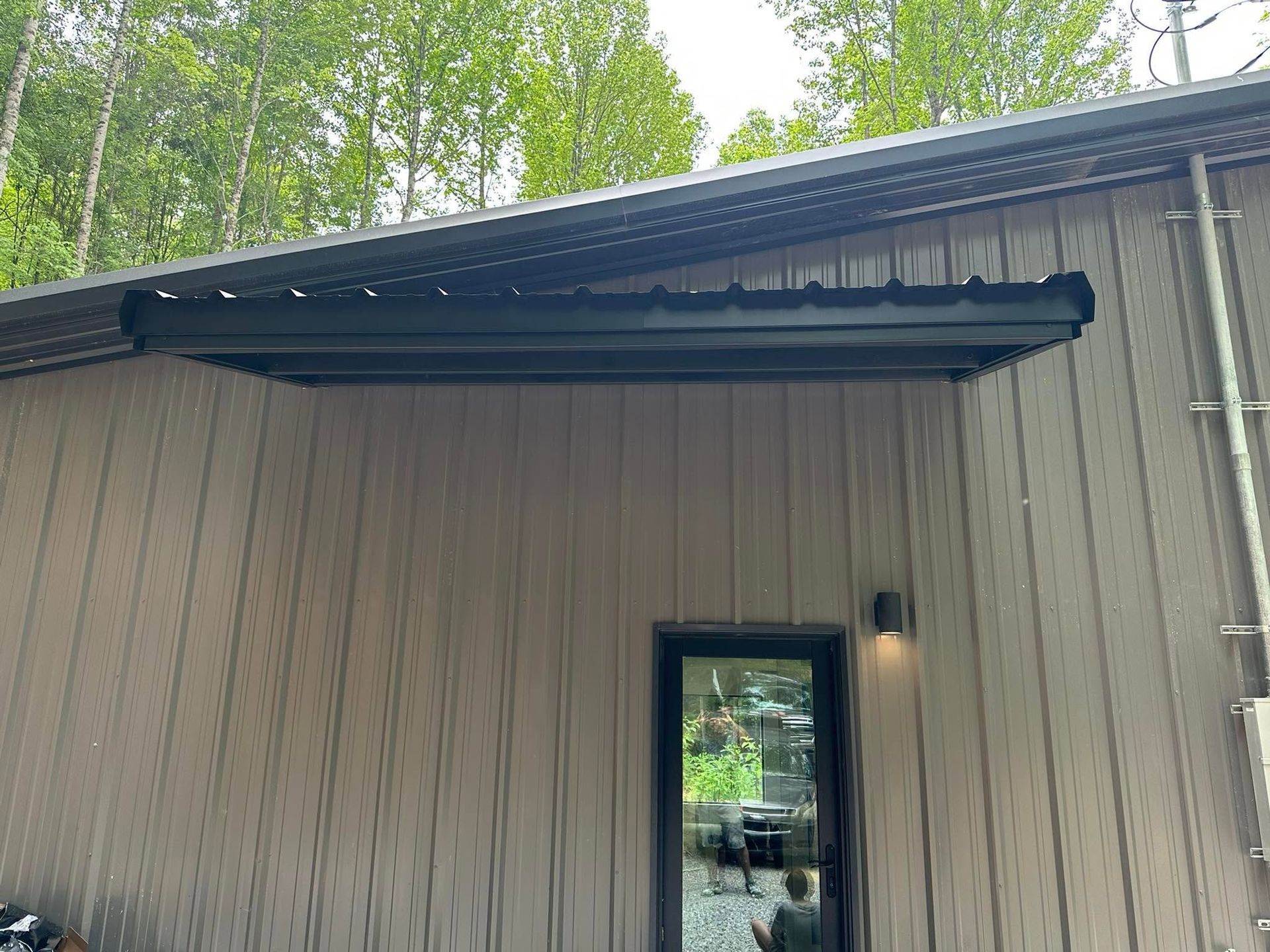 Exterior of a building with a dark awning, tan siding, and a glass door under trees