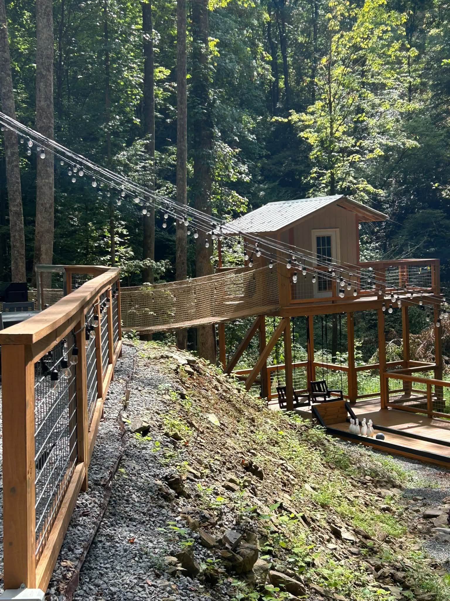 Wooden suspension bridge in a forest, leading to a small cabin among tall trees.