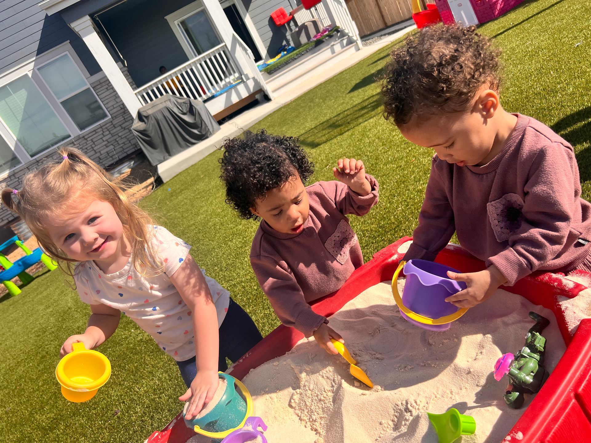 Three toddlers playing in a sandbox with colorful toys on a sunny day.