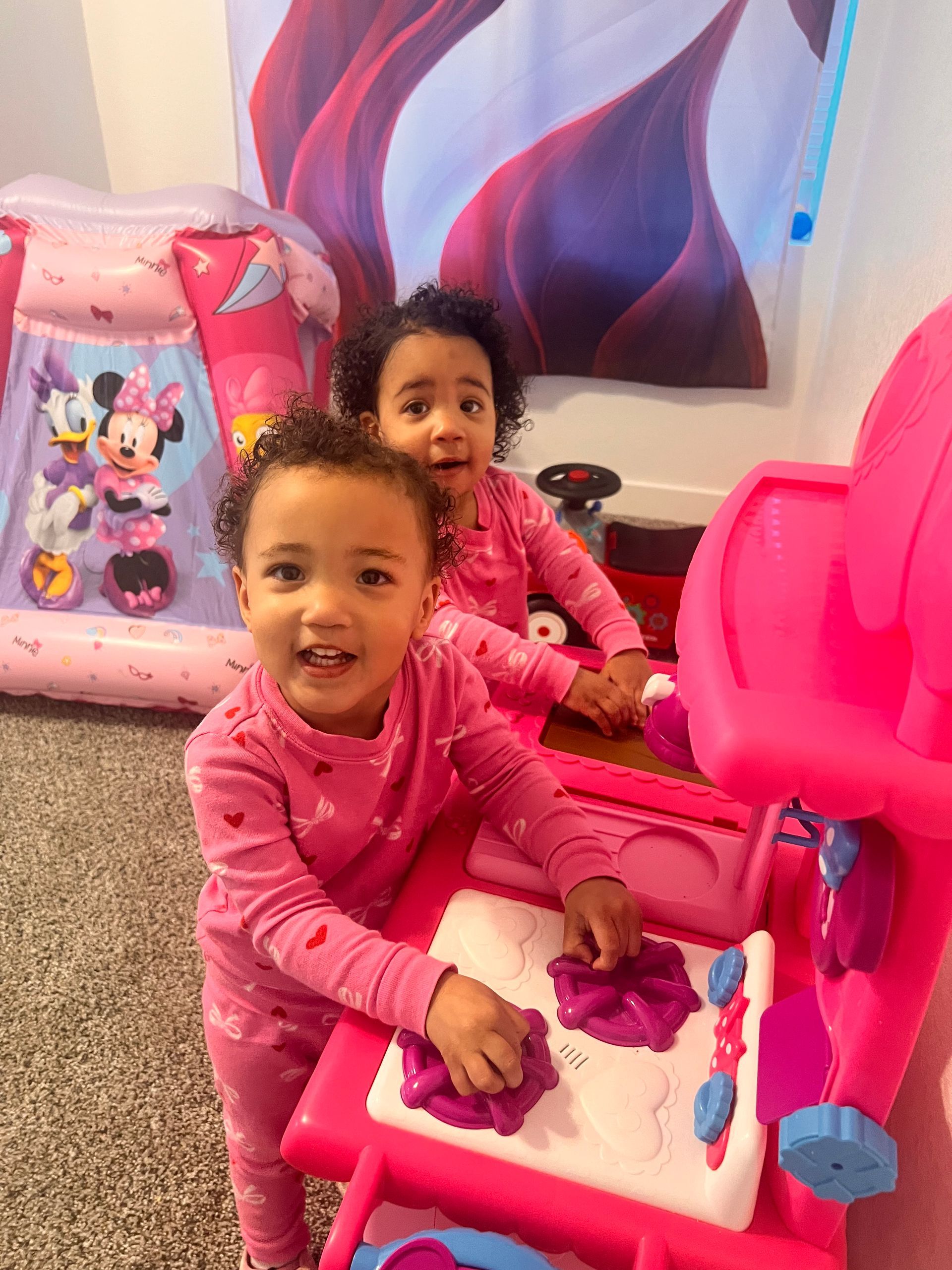 Two children in pink pajamas playing with a pink toy kitchen, smiling.