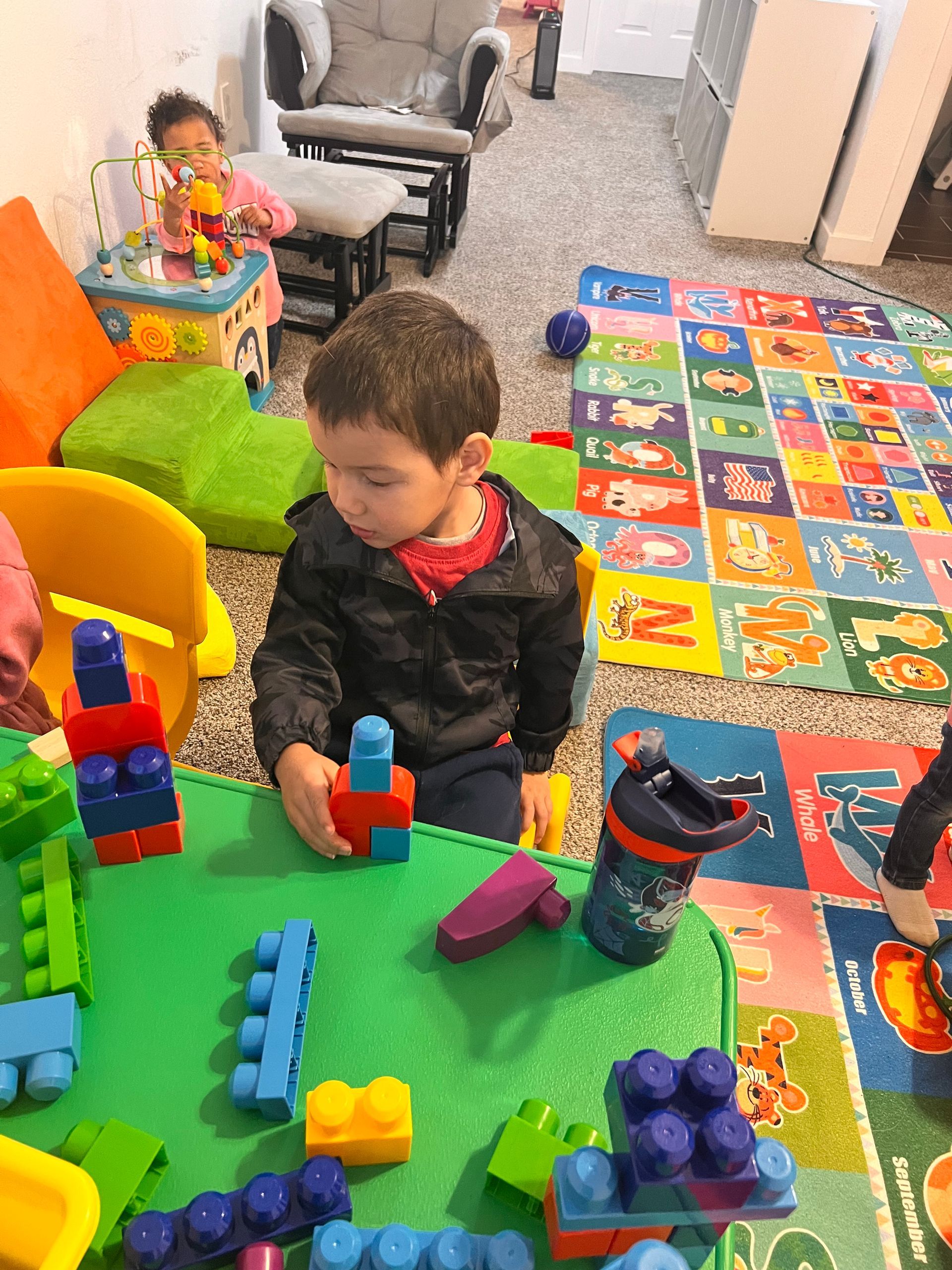 Child playing with blocks on a green table, another child in the background. Playroom setting, colorful toys.