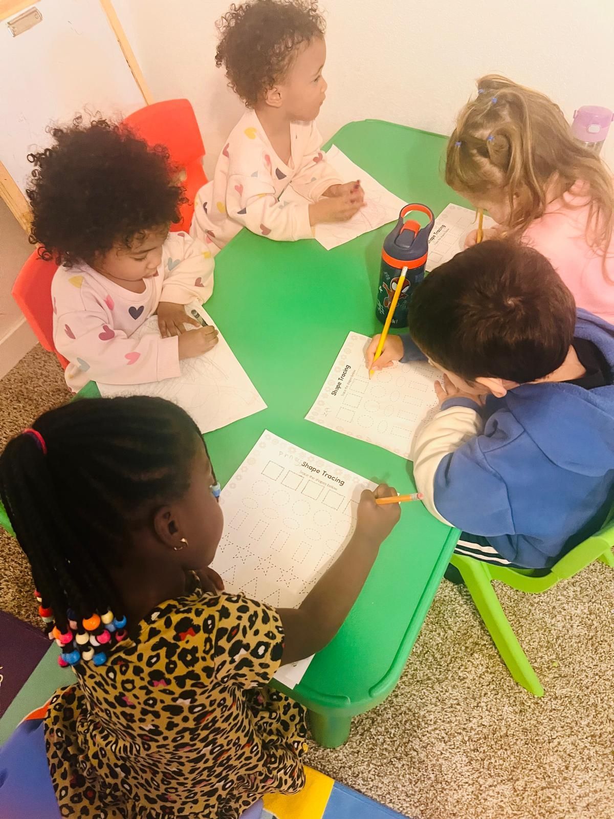 Children seated around a green table, drawing on paper with pencils.