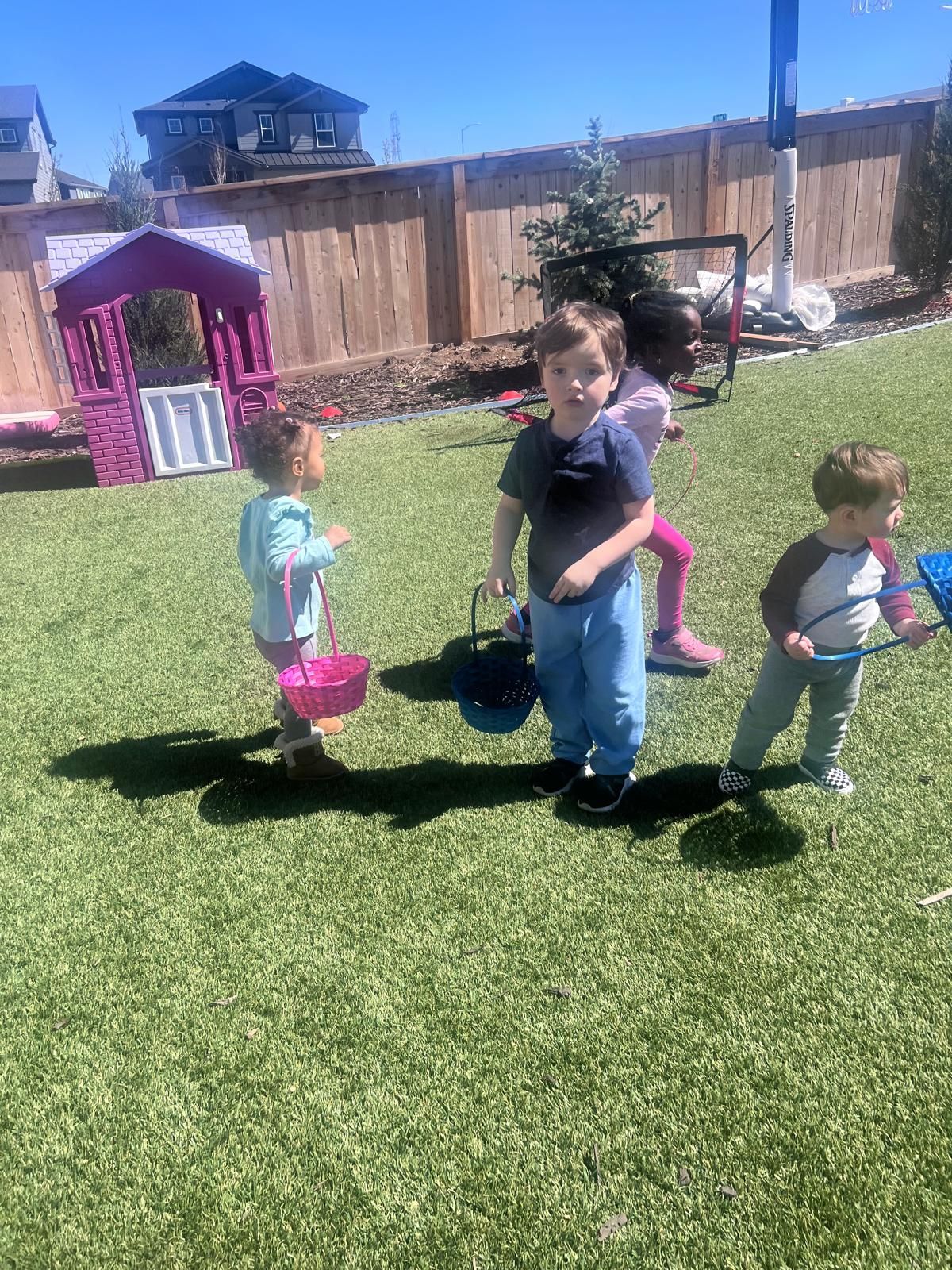Three children on a sunny lawn carrying buckets, with a playhouse and fence in the backyard.
