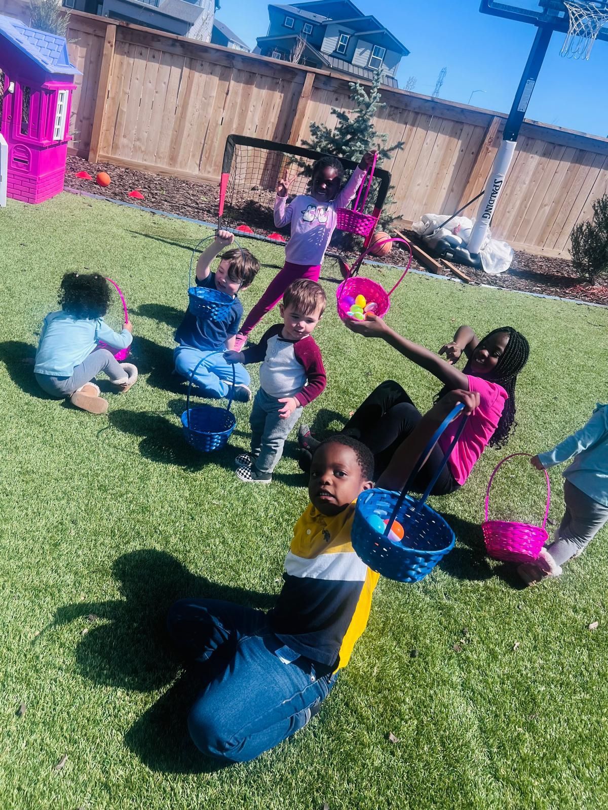 Children sitting in a grassy yard with pink baskets, facing a person standing near a wooden fence.