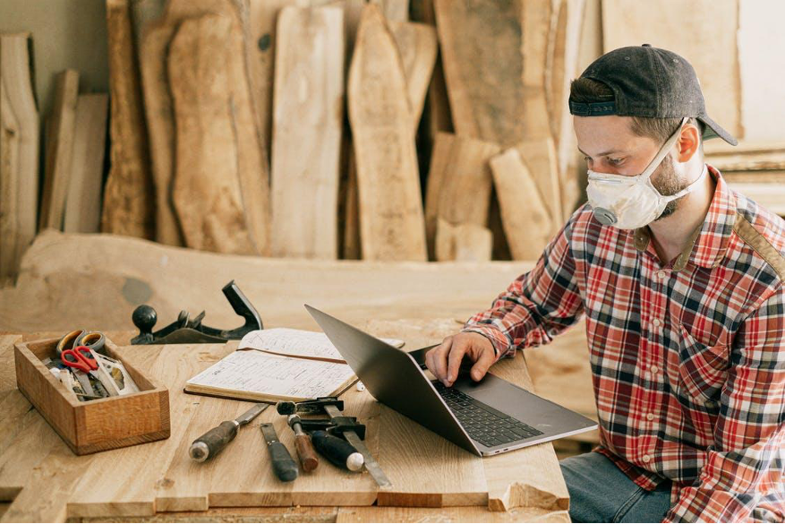 Woodworker with mask and cap working on a laptop at a workbench with tools and lumber in the background.