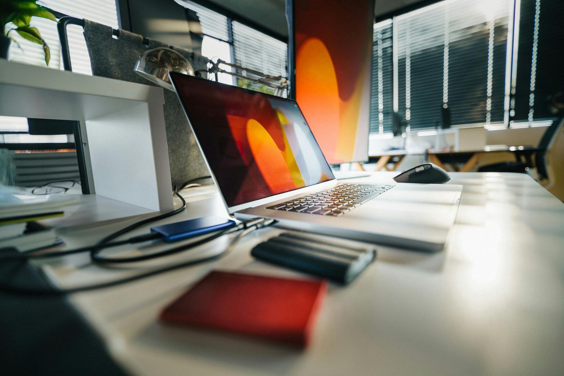 A laptop sits open on a bright, cluttered desk next to a mouse, a small blue drive, and a red notebook.