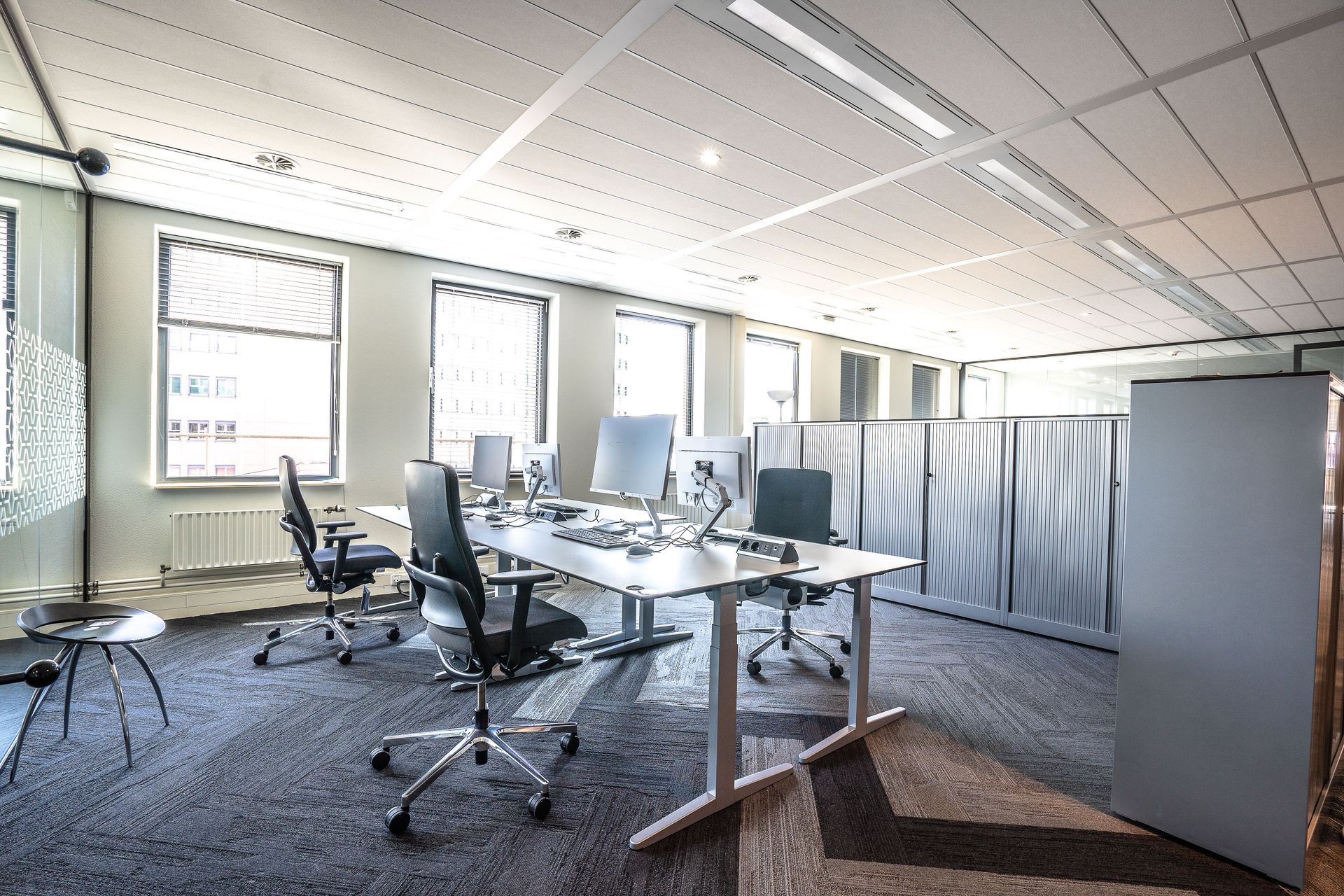 Modern office with desks, swivel chairs, monitors, and gray carpet under bright windows and ceiling lights