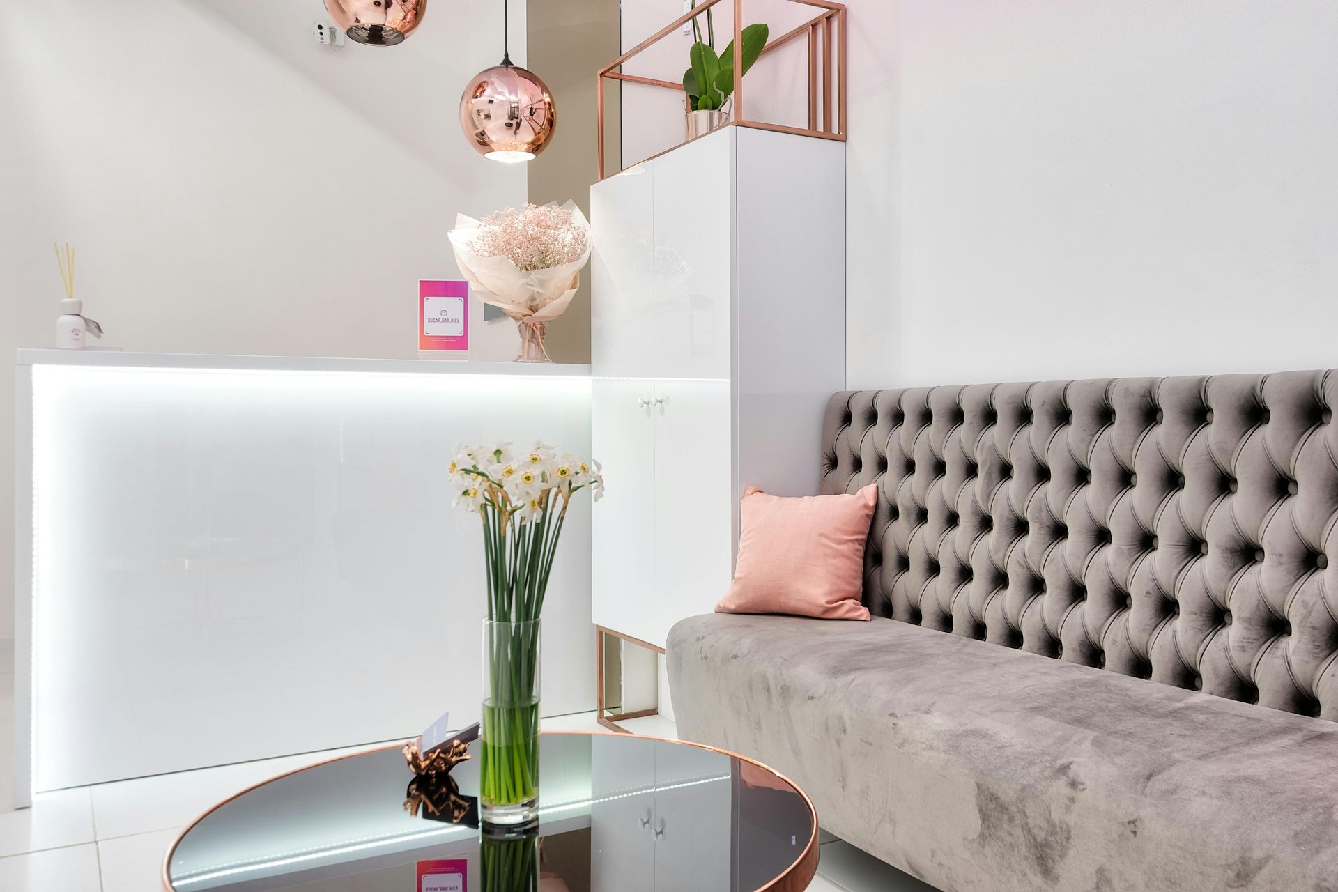 Modern reception area with gray tufted sofa, white cabinet, glass table, and pink accents