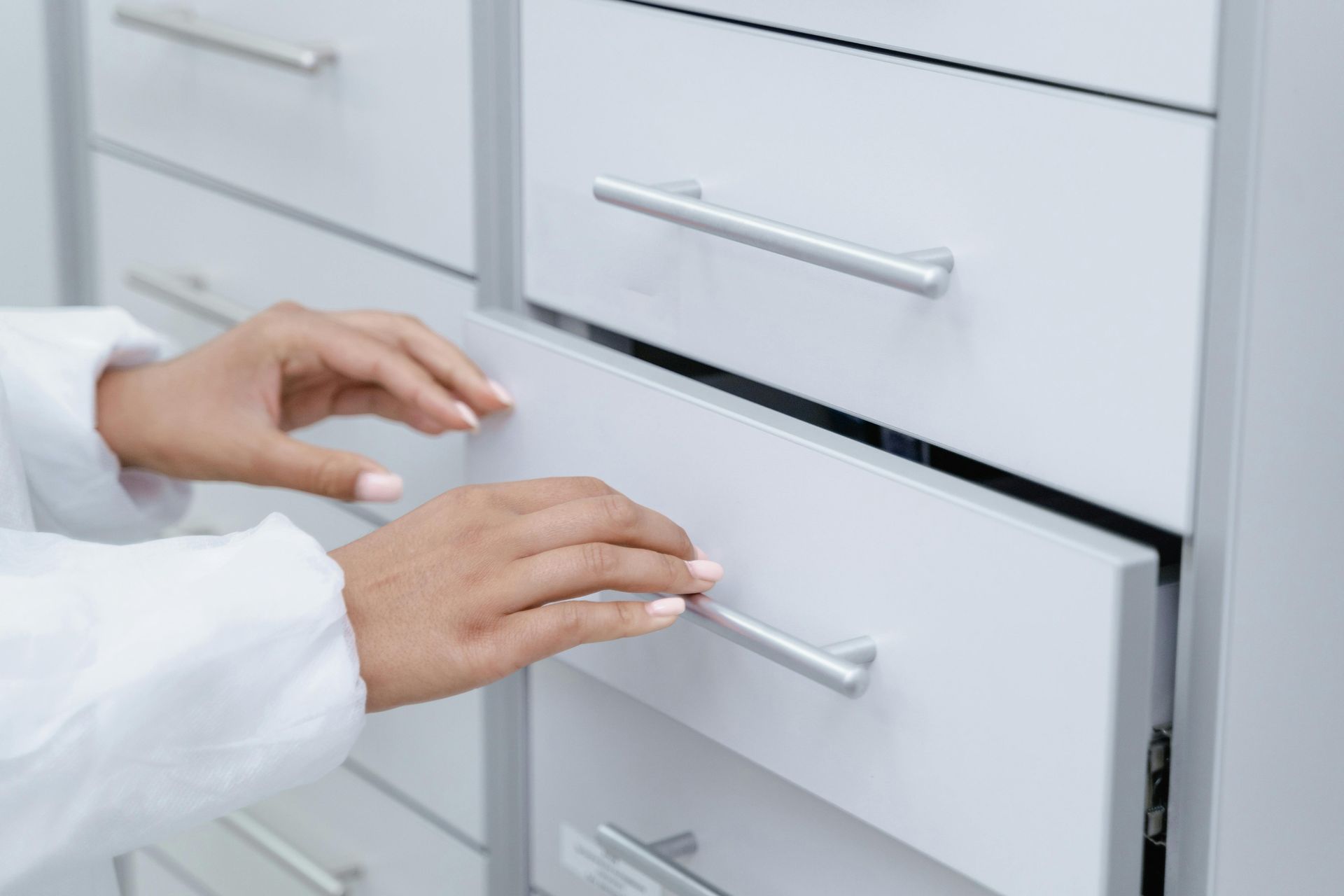 Hands opening a white cabinet drawer with metal handles in a bright kitchen or office.