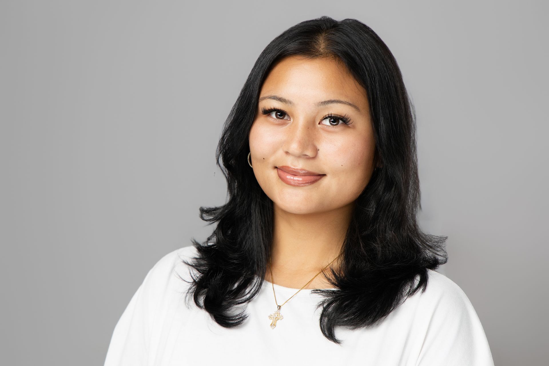 Portrait of a woman with shoulder-length black hair wearing a white top, smiling against a gray background
