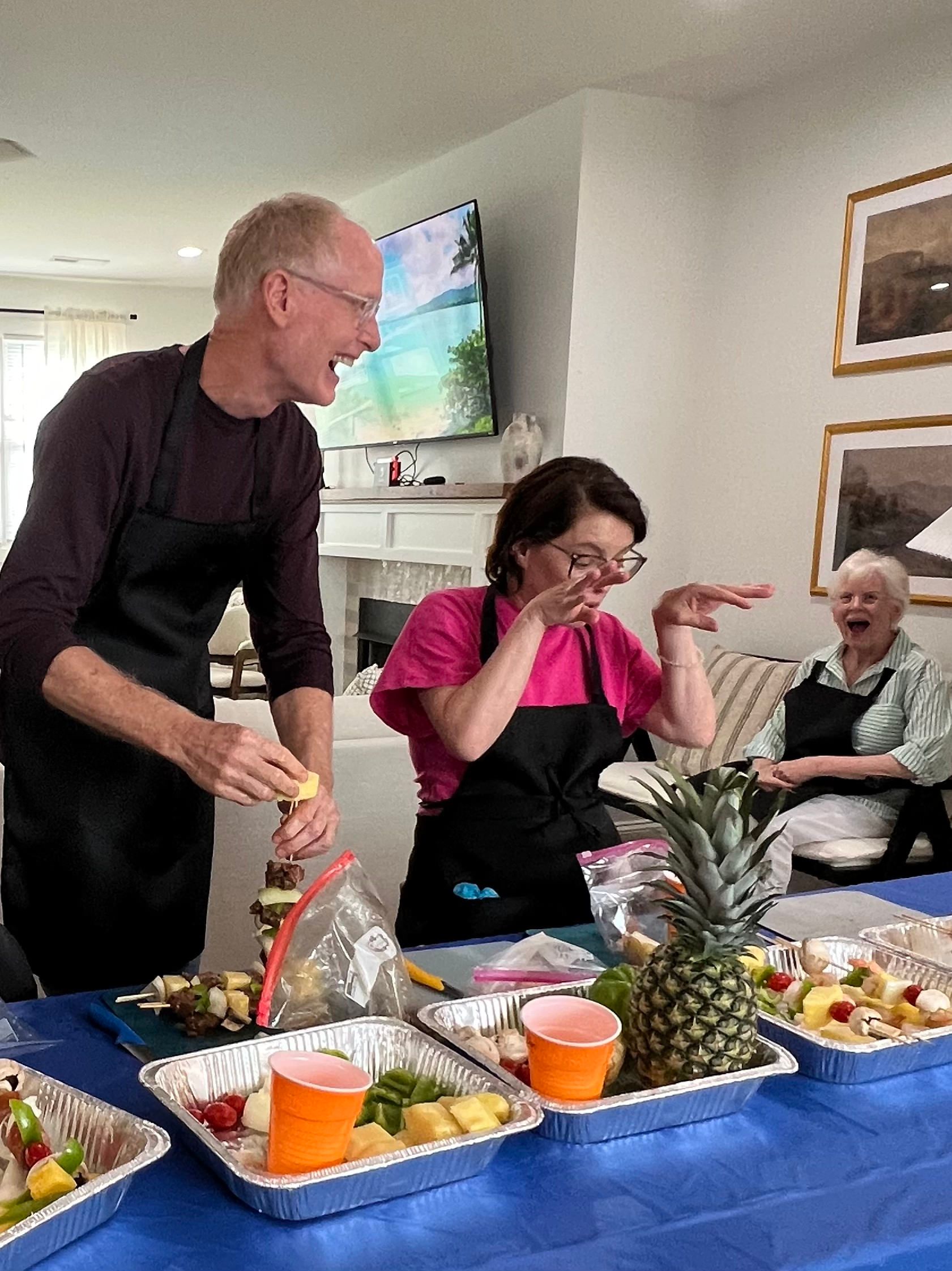 People preparing fruit skewers, laughing, at a table with food, pineapple, and cups.