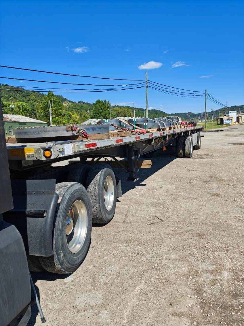 Flatbed trailer loaded with metal beams parked on gravel under a blue sky