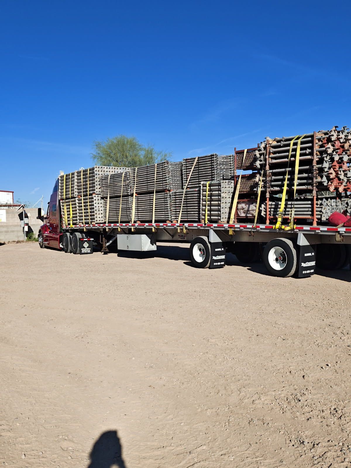 Flatbed truck loaded with stacked pallets parked on a dirt lot under a clear blue sky